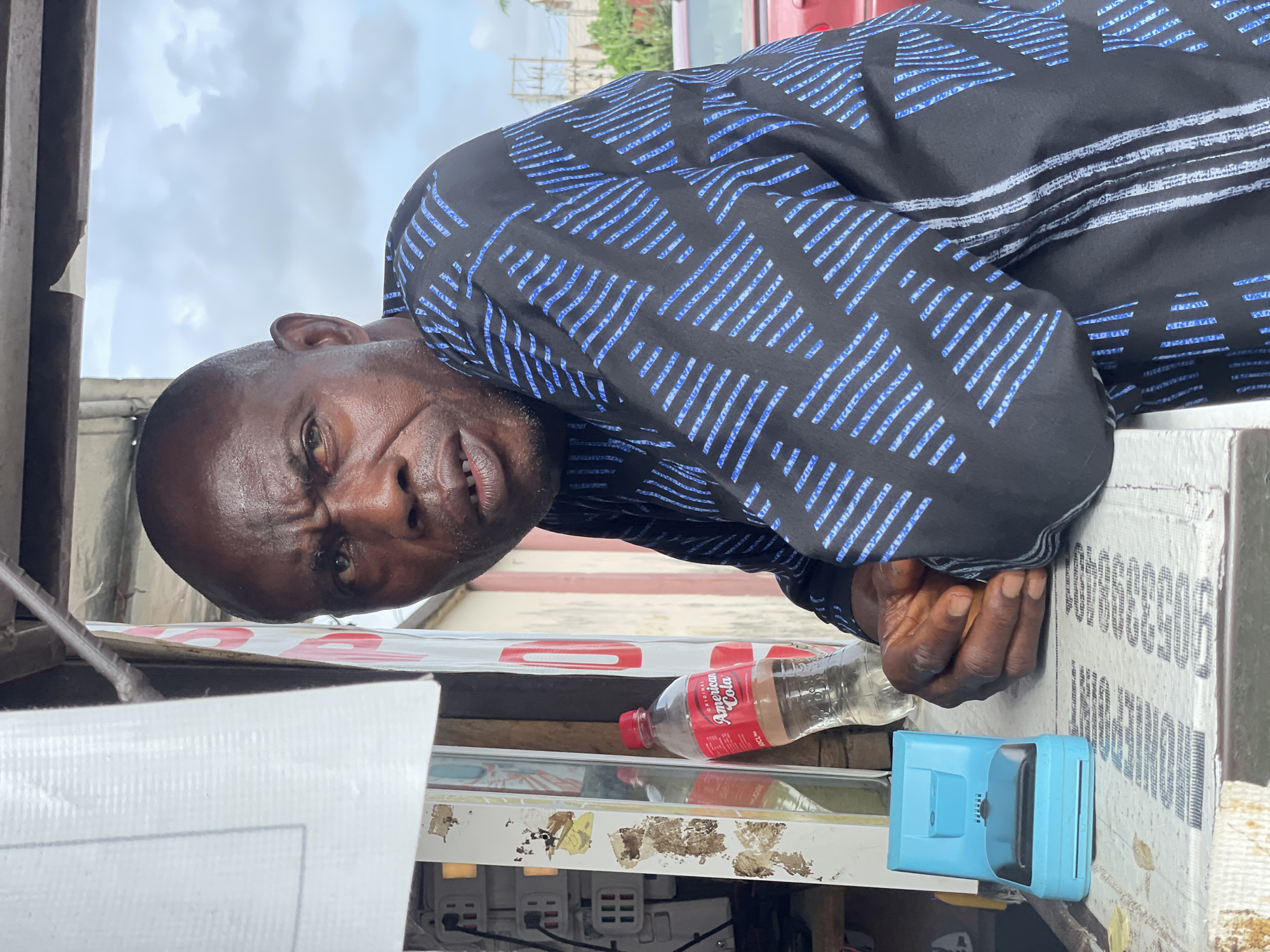 A middle-aged, dark-skinned man in a black and blue patterned shirt is leaning over a white counter in Ado, Nigeria. He is looking directly at the camera with a slight frown. On the counter, there is an almost empty bottle of "American Cola," a light blue credit card terminal, and electrical outlets. The background includes a building and a partially visible red vehicle.