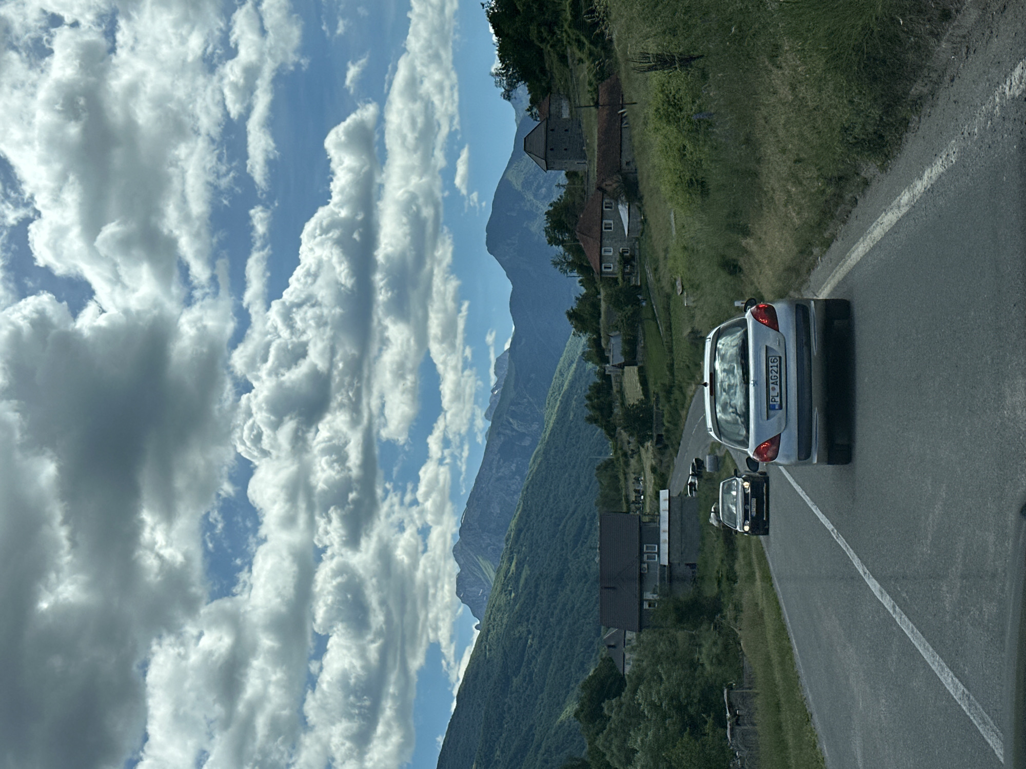A wide shot captures a scenic drive through a rural mountainous region under a dynamic sky filled with fluffy white and gray clouds. The road, asphalt with a white line marking the lanes, curves through a landscape dotted with houses and lush green hills. In the foreground, two cars are traveling along the road; a silver hatchback is in the right lane, and a darker car is in the left lane. In the background, majestic mountains rise, their slopes covered in dense green vegetation, with some jagged peaks visible against the sky. Several houses, with red or dark roofs, are nestled into the hillsides, some appearing to be older stone structures and others more modern. Trees and bushes fill the spaces between the houses and along the roadside, creating a vibrant, natural environment. The overall impression is one of a peaceful journey through a beautiful, undeveloped area.