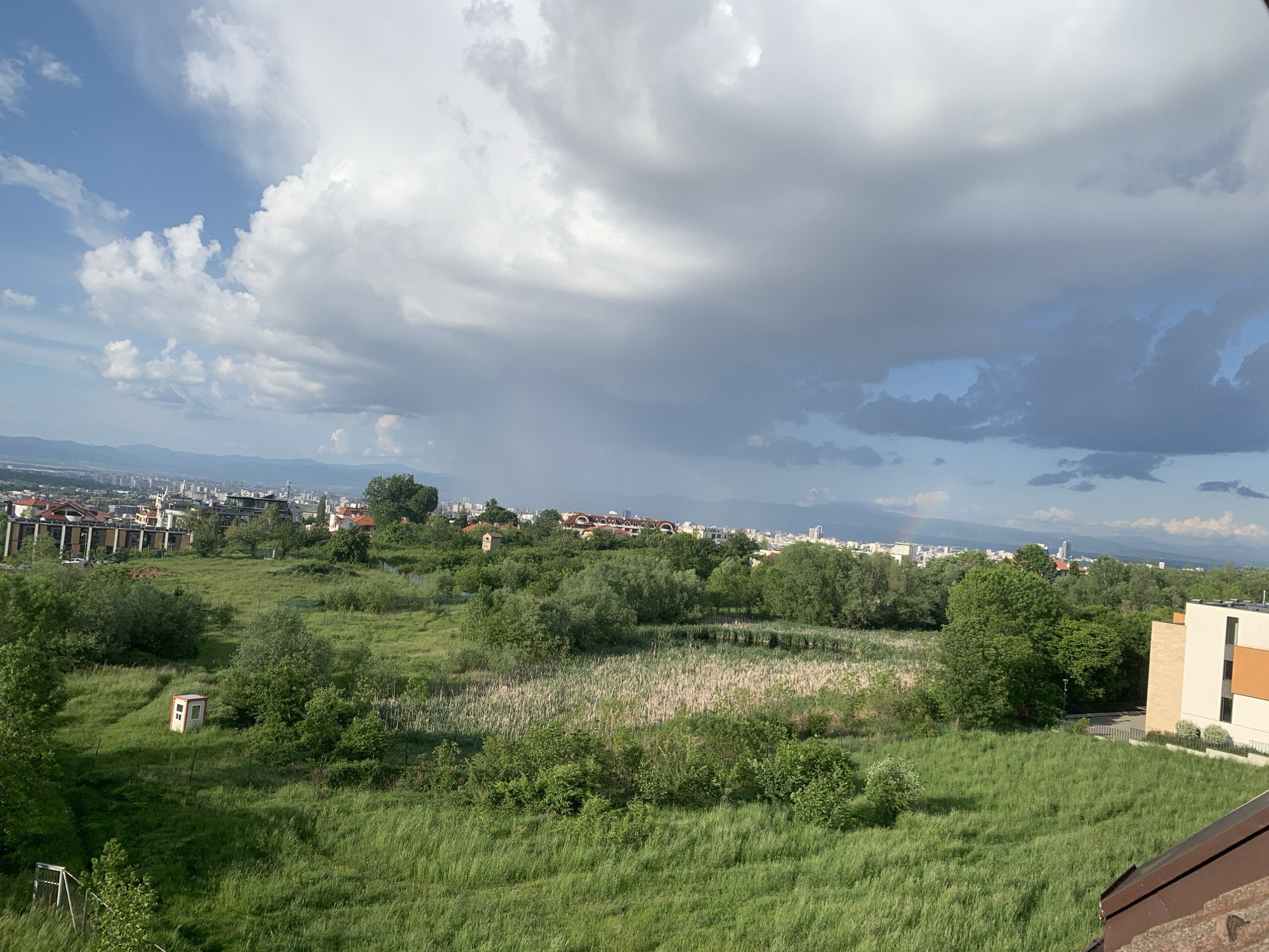 The image shows a cityscape of Unknown, Unknown, with a large green field in the foreground. In the middle distance, a cluster of buildings and trees separates the field from the city skyline in the background. The sky is predominantly cloudy with patches of blue sky, and a faint rainbow is visible in the distance. A small, shed-like structure stands in the middle of the field. The light indicates it is daytime.