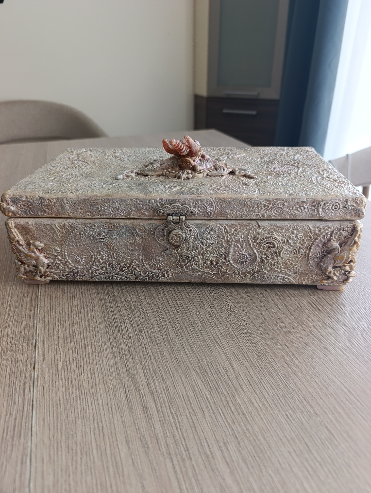 A closed, rectangular decorative box with ornate, textured detailing sits on a light-colored wooden surface. The box is a muted metallic color, possibly bronze or aged gold, with intricate patterns carved into its lid and sides. The lid is adorned with a raised, reddish-brown embellishment resembling a shell or a coiled insect. A small, decorative latch secures the front of the box. The background is blurred, showing a sliver of a chair and a tall cabinet with dark wood and frosted glass doors, suggesting an indoor setting, possibly a home or office. The lighting is soft and diffused. There is no indication of the city of Toruń, Poland, or any specific forensic details related to a crime scene.