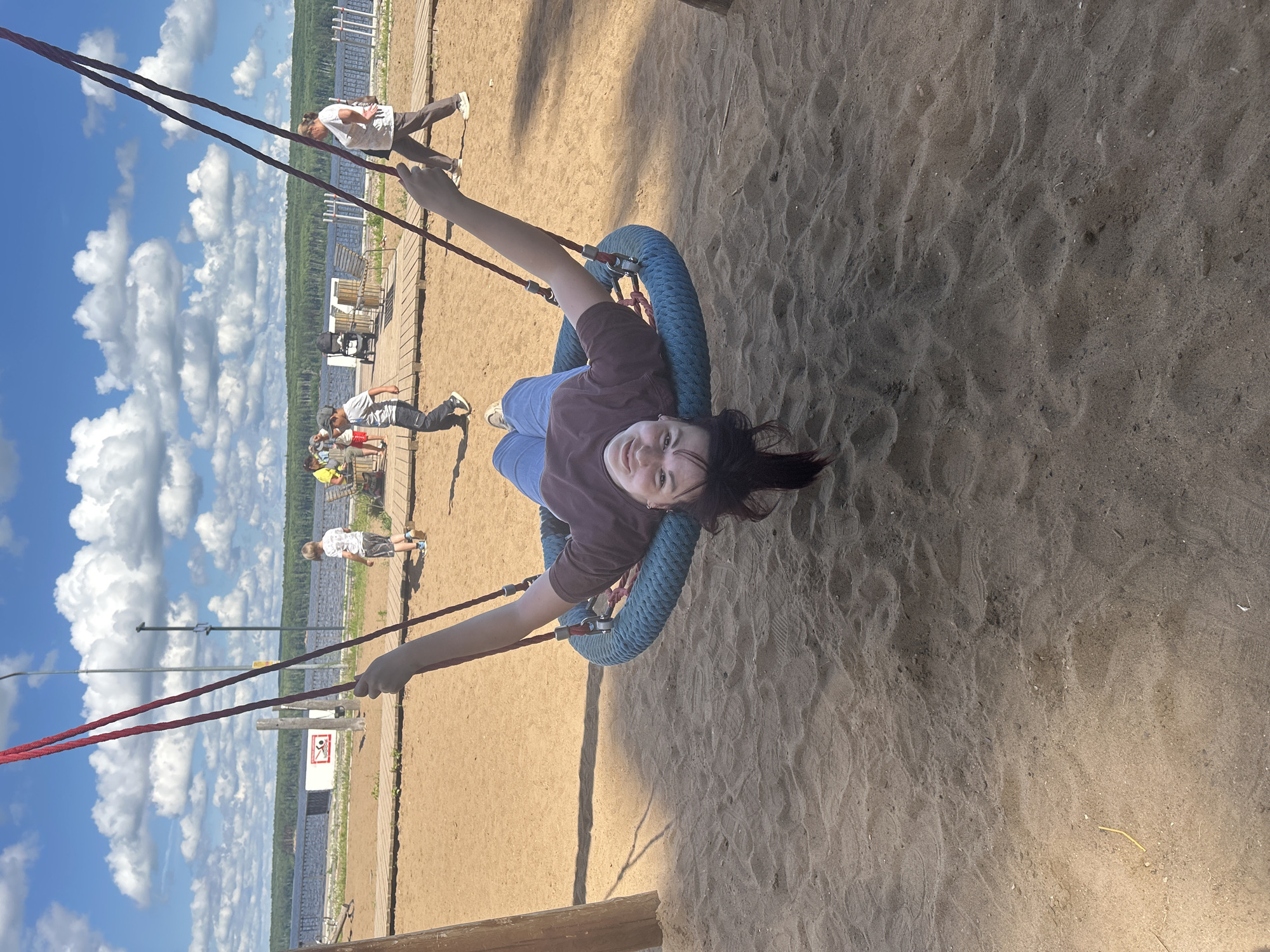 In Chaikovsky, Russia, a young woman with dark hair lies on her back on a large, circular rope swing at a sandy playground. She is wearing a brown t-shirt and blue jeans, and her legs are extended upwards. The swing is suspended by two thick red ropes from a structure that is partially visible. The background shows a body of water with a forested shoreline under a bright blue sky with scattered white clouds. Several other people, including children and adults, are visible in the background, some walking on a wooden pathway near the water. The scene appears to be a sunny day in a recreational area.