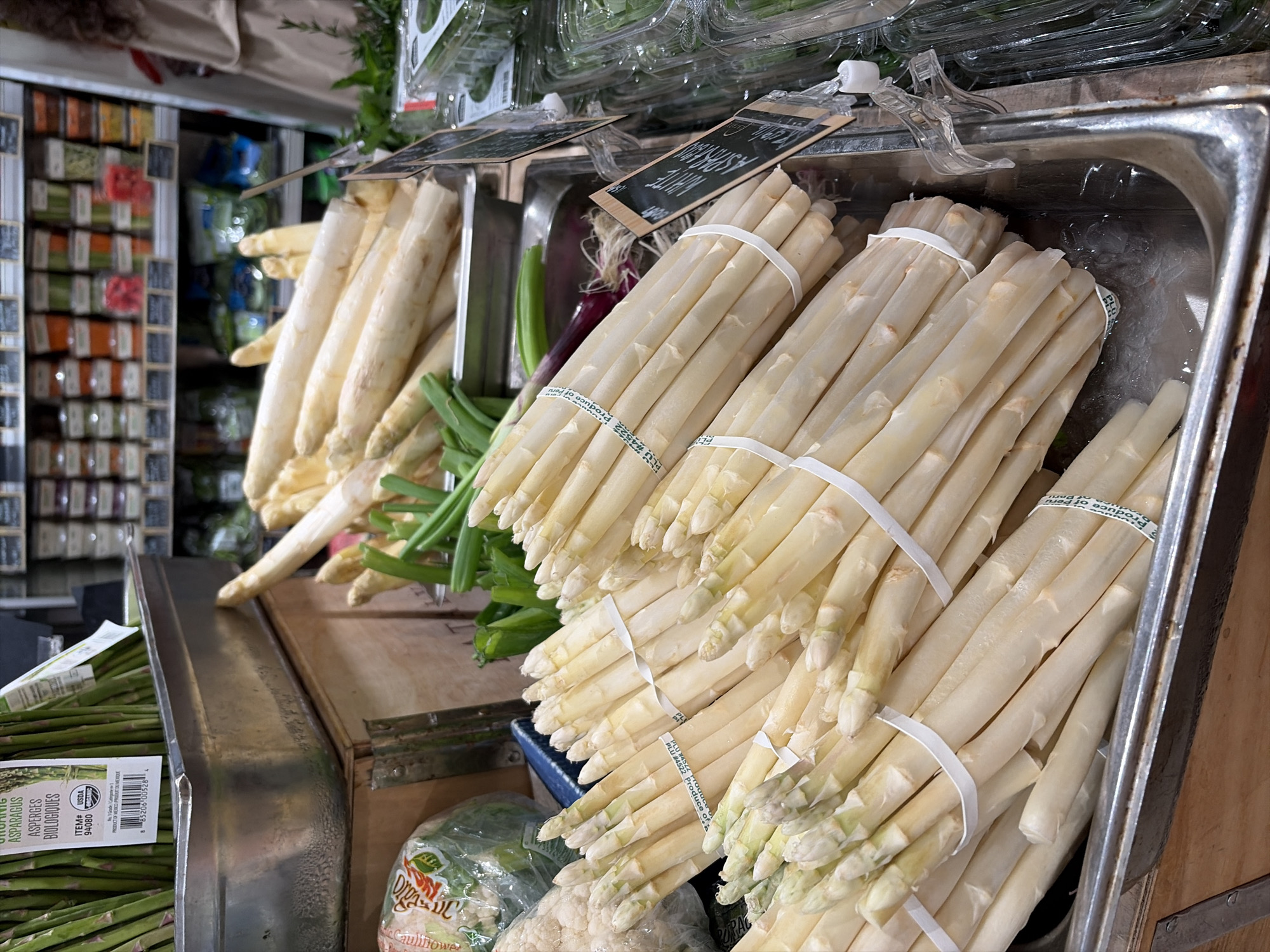 The image shows a close-up view of bundles of white asparagus in a metal display bin filled with ice. The asparagus is tied with white bands and some bundles have PLU stickers attached. In the background, there are shelves stocked with various produce items, including green asparagus, cauliflower, and packaged greens. There are also price tags visible on some of the produce. The setting appears to be a grocery store or market. The image does not contain any explicit information about the location in New York City, United States of America, or any specific people or actions.