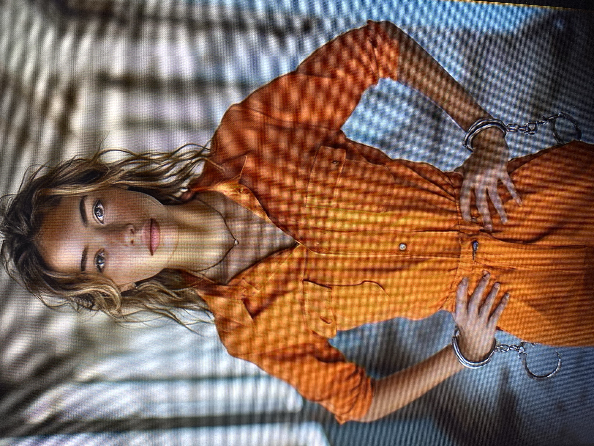 A young woman with wavy blonde hair, wearing an orange jumpsuit and handcuffs, stands with her hands on her hips in a dimly lit hallway. The hallway appears to be part of an industrial or correctional facility, with concrete walls and overhead lighting. The woman's gaze is directed at the camera, and her expression is neutral. The background is blurred, but suggests a long corridor with repetitive architectural features.