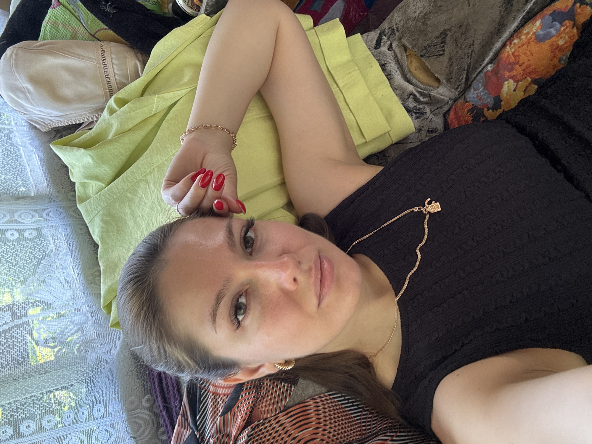 A woman with brown hair and brown eyes, wearing a black sleeveless top and a gold necklace with a charm, is lying down. Her right hand, with bright red nail polish, is resting on her forehead, and she is wearing a gold bracelet. Behind her, there are piles of clothing, including a lime green jacket and other garments. A window with white lace curtains is visible in the background, suggesting an indoor setting. The overall lighting is natural. The exact location in the city of Unknown, Unknown cannot be determined from the image.