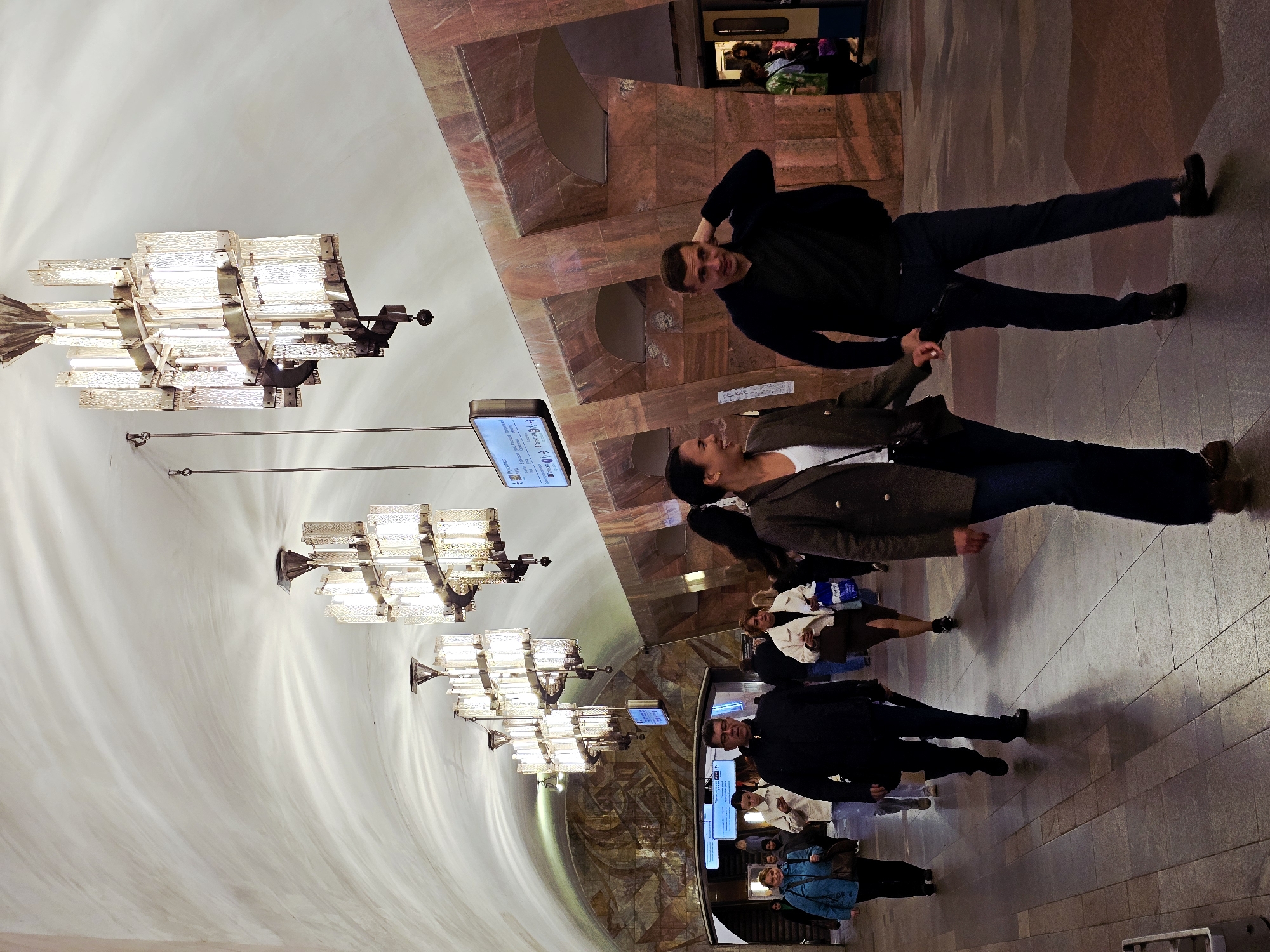 The image depicts a scene within a Moscow Metro station in Russia. Numerous people are visible, mostly walking through the station. A man and woman are positioned towards the upper part of the image, standing and holding hands. The man is wearing dark clothing, while the woman is wearing a blazer. The station features large chandeliers hanging from a curved, high ceiling. The walls are decorated with reddish-brown marble. Signs with train schedules are hanging from the ceiling.