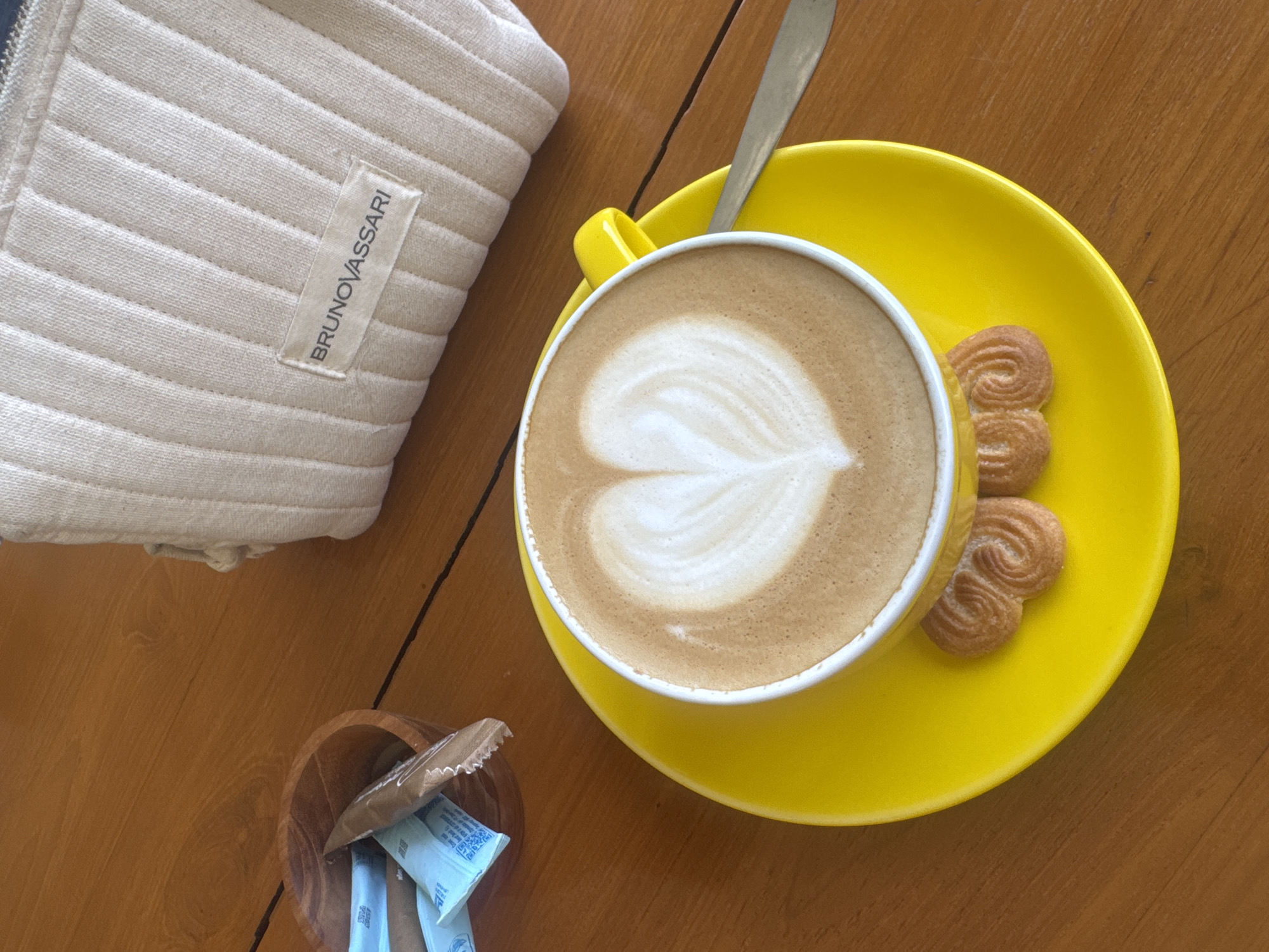 The scene is a top-down view of a table setting. A yellow cup filled with a latte, featuring latte art in the shape of a heart, rests on a matching yellow saucer. Two heart-shaped cookies are placed on the saucer next to the cup. A silver spoon is positioned in the cup. To the left of the cup is a beige, quilted item with a "BRUNOVASSARI" label. A small wooden bowl containing sugar packets is in the lower-left corner. The table appears to be made of wood. The location is Kuta Selatan, Indonesia.