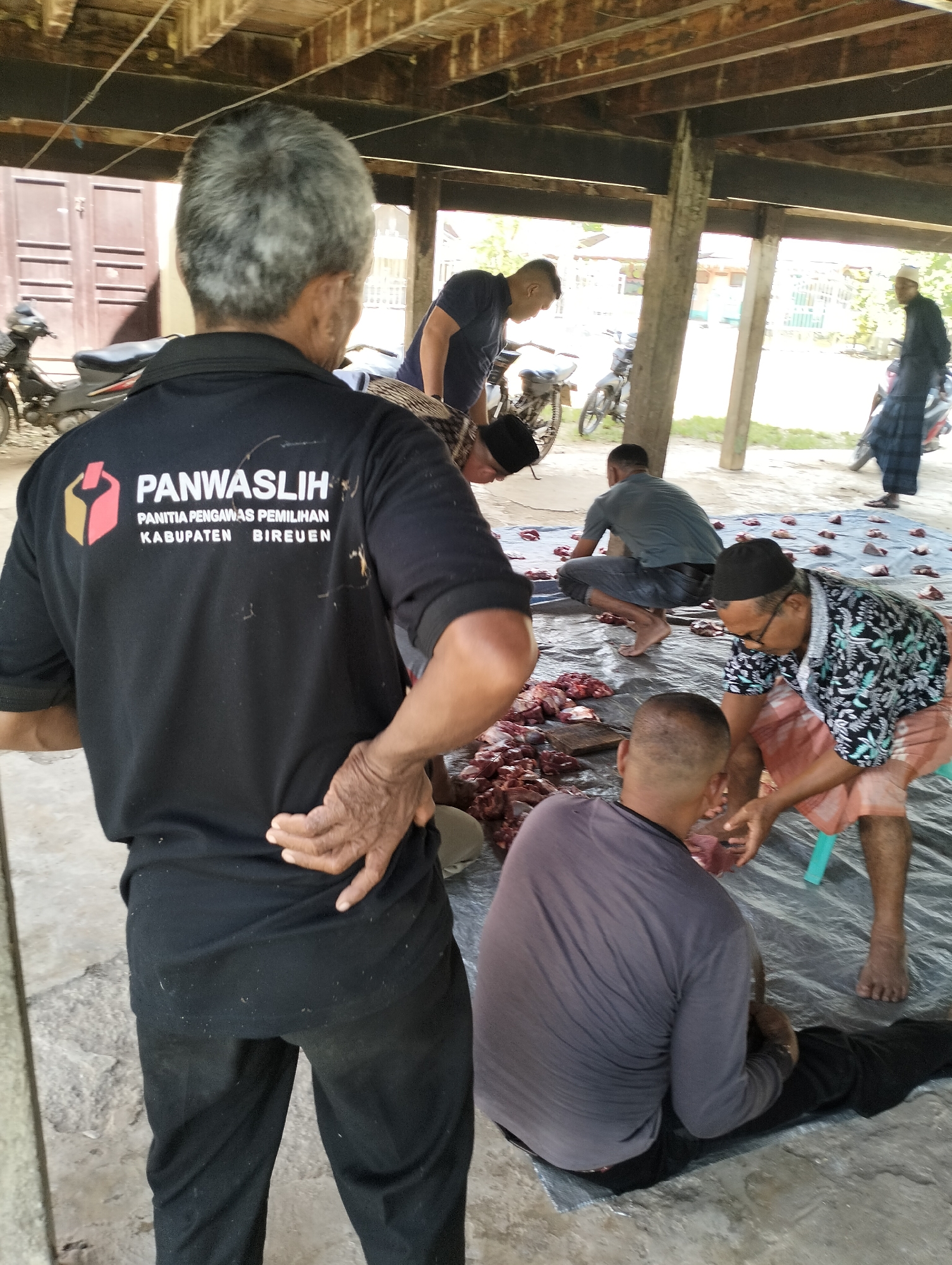 The image captures a scene of several men engaged in butchering meat, likely beef or lamb, outdoors under a wooden structure in Meunasahalue, Indonesia. A group of men are working on a sheet of blue plastic laid on the ground. The meat has been divided into smaller pieces. The men are sitting or squatting around the pile of meat, cutting and preparing it for distribution. One man stands with his back to the camera wearing a black shirt with the Indonesian words "PANWASLIH" and "PANITIA PENGAWAS PEMILIHAN KABUPATEN BIREUEN." Several motorcycles are parked in the background.