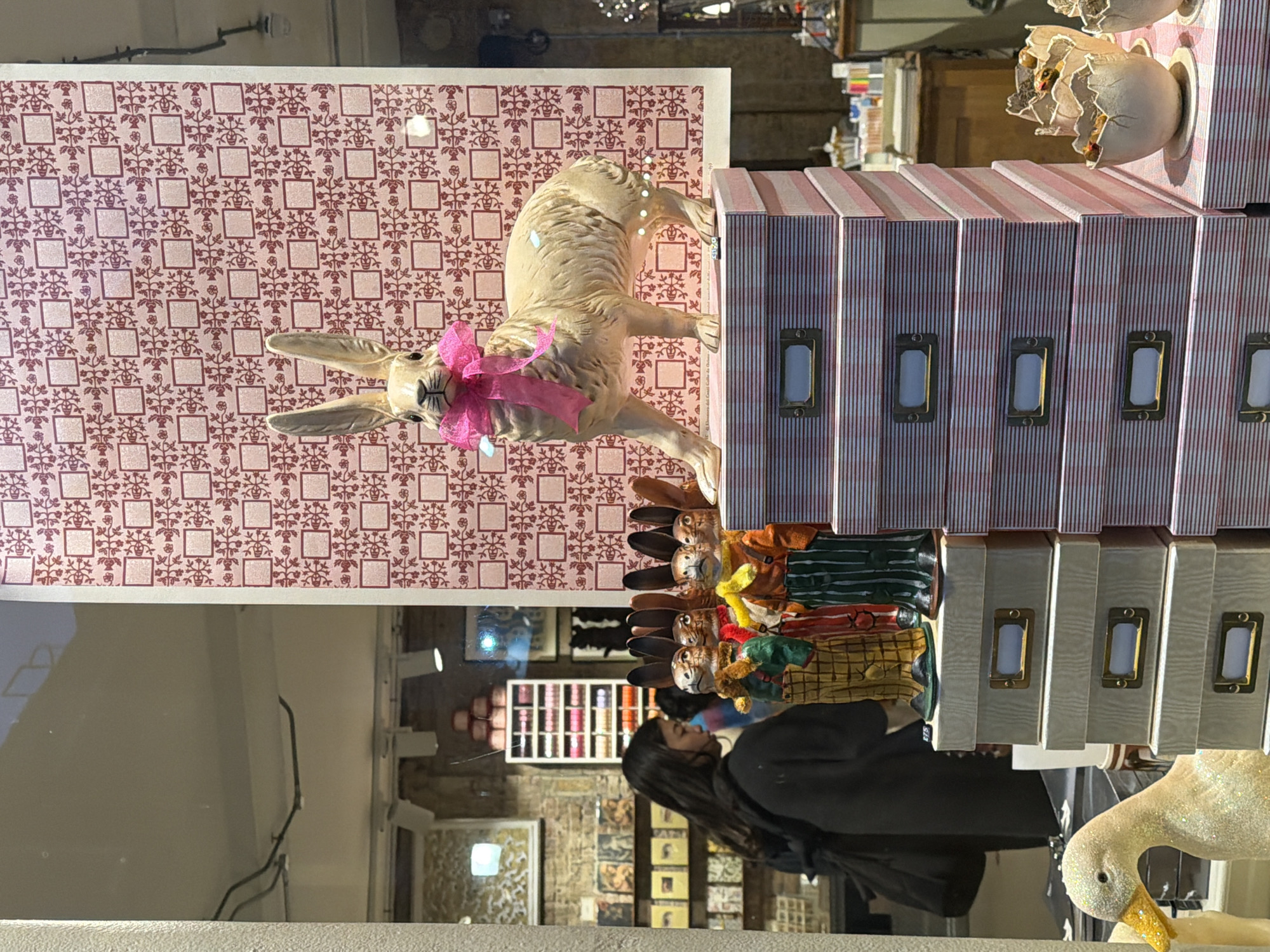 The image depicts a retail shop window display, observed from the exterior, looking into the store's interior in an urban environment in London, United Kingdom.
In the foreground, a large, cream-colored rabbit statue, featuring prominent ears and a bright pink ribbon tied around its neck, stands on a stack of light pink and white striped rectangular boxes. To the left of this, on a separate stack of light grey boxes with brass-colored label holders, are three smaller, anthropomorphic rabbit figurines dressed in colorful garments. Further left, partially visible, is a white, glitter-covered duck figurine with a yellow beak. To the right, on a lower stack of striped boxes, several white, cracked eggshell-shaped containers each hold small chick figurines. The background of the window display features a large sheet of pink wallpaper adorned with a repeating pattern of red floral motifs and square outlines.
Through the window, the shop's interior is visible. A young adult female with long dark hair, wearing a dark top and carrying a dark shoulder bag, stands facing away from the viewer, appearing to browse merchandise on shelves. Behind her, shelves are stocked with what appear to be books, cards, or boxed items. A tall, narrow white shelving unit displaying numerous spools of multicolored thread or ribbon is also visible. The store's interior features exposed brick walls and ceiling-mounted spotlights, characteristic of a boutique or specialty store in a commercial district. The absence of specific street names or identifiable landmarks prevents pinpointing the precise address, but the architectural style and retail context are consistent with various shopping areas throughout London.