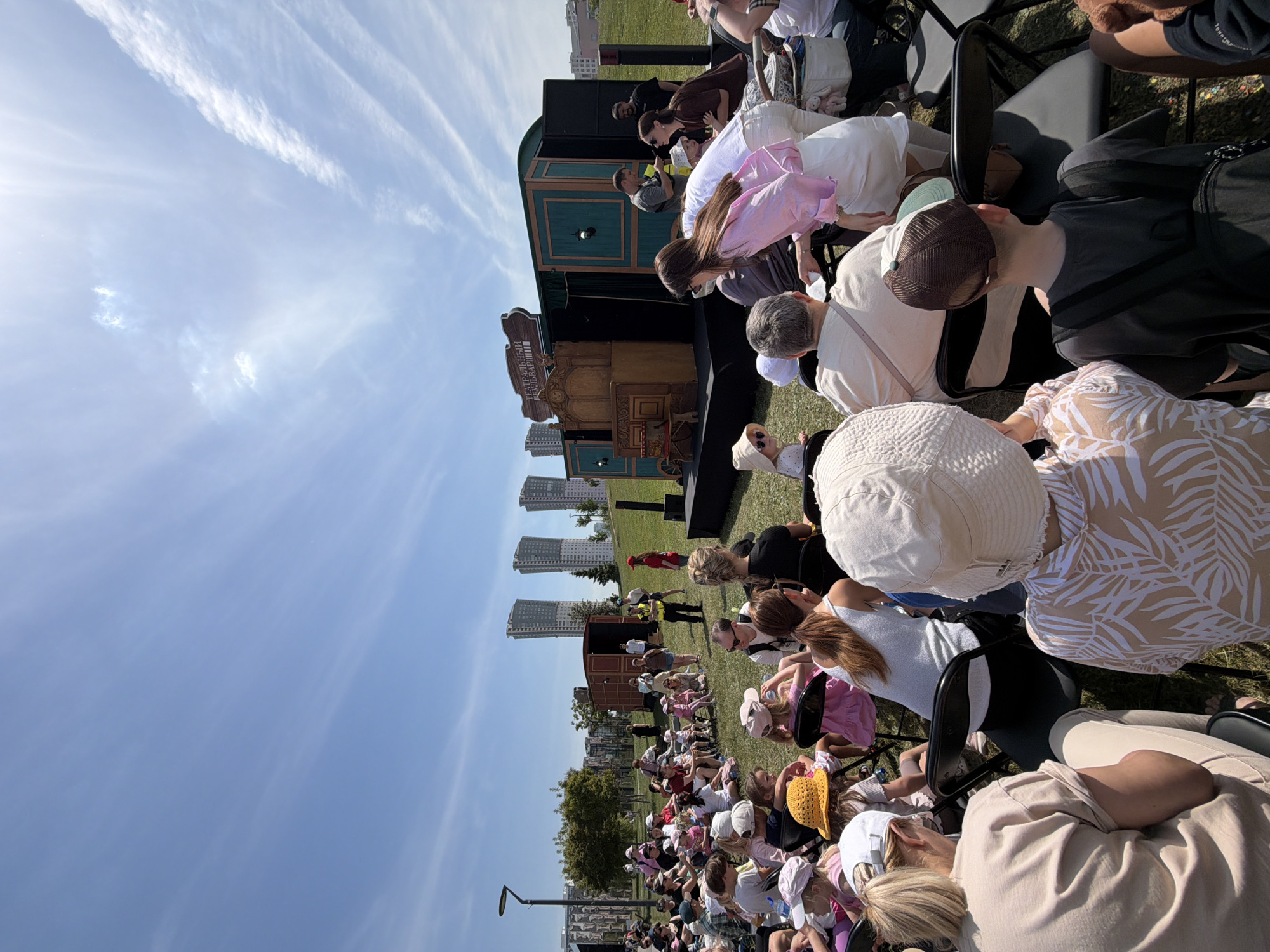 This detailed image captures an outdoor theatrical event in Khimki, Russia, on a bright, sunny day. The scene is set in a green grassy area, likely a park, with several modern high-rise residential buildings visible in the background under a clear blue sky with sparse wispy clouds.
In the foreground, numerous individuals, primarily adults and children, are seated on black folding chairs and directly on the grass, forming an audience. Most are facing towards a portable stage setup in the middle ground. Many audience members wear hats or caps for sun protection, and their attire consists of casual summer clothing. Visible in the immediate foreground, facing away from the camera, is a person in a white wide-brimmed hat and a light-colored shirt with a tropical leaf pattern. Next to them, a child with a brown and light green baseball cap is also seated, facing the stage. A woman in a light pink long-sleeved shirt and white skirt stands near the center of the frame, observing the stage.
The stage itself is designed with elements resembling old-fashioned train cars or wagons, painted in dark green and brown tones. A large wooden structure, possibly an ornate organ or cabinet, is positioned centrally on the stage. Above this structure, a sign reads "ТЕАТРАЛЬНЫЙ БУЛЬВАРЪ" (Theatre Boulevard) in Cyrillic. Several individuals are present on or immediately around the stage. One person wears a yellow safety vest over a dark uniform, standing near the stage's edge, possibly an event organizer or security. Another person in a red shirt is also seen near the stage. To the far left, a modern street light post stands tall. The overall atmosphere suggests a community cultural event taking place under pleasant weather conditions.