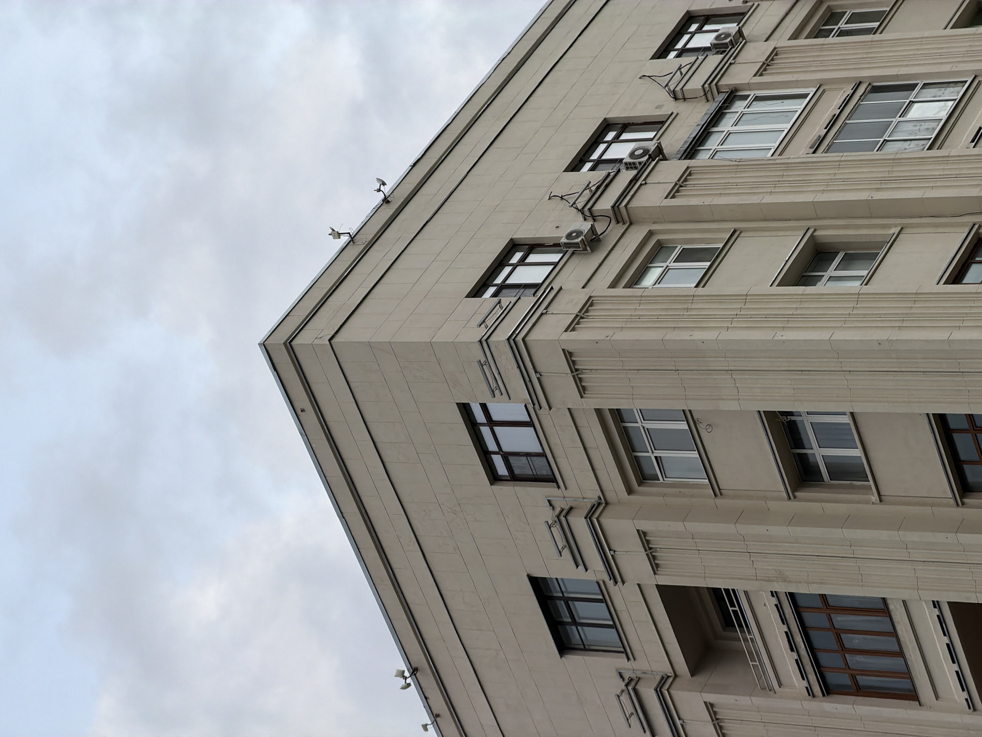 The image shows the facade of a building with a light beige color. The building has multiple floors with numerous windows. Some windows are dark-framed and reflect the sky, while others are lighter. There are architectural details on the facade, including vertical fluting and projecting elements. Air conditioning units are visible mounted on some of the walls. On the roofline, there are a few small structures that appear to be antennas or surveillance cameras. The sky is overcast with a muted grey color. The image appears to be taken from a low angle, looking upwards at the building. The context provided indicates this is likely in Moscow, Russia.