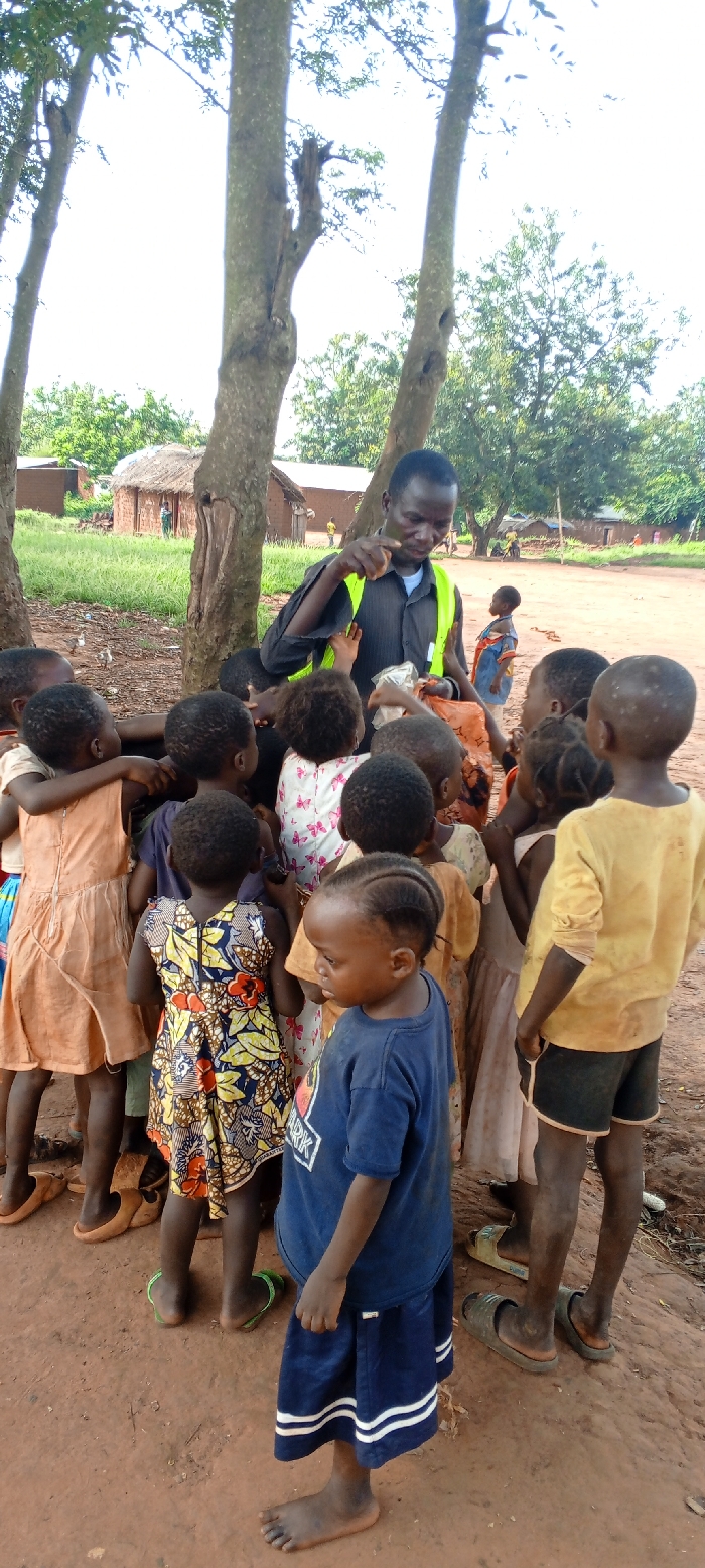 The image depicts a man interacting with approximately ten young children in an outdoor, rural setting in Ngalula, Democratic Republic of the Congo.
**People:**
* **The Man:** Positioned centrally, facing the children, slightly to the left of the frame's center. He is an adult male with dark skin and short, dark hair. He wears a dark, possibly black or navy, collared button-up shirt with short sleeves and a bright neon yellow-green reflective vest over it. His right arm is bent at the elbow, hand extended with fingers pinched together, possibly distributing something. His left hand holds a transparent plastic bag containing a light-colored substance and a larger orange bag or pouch. He appears focused on the children.
* **The Children:** A group of approximately ten children, ranging from toddlers to early elementary age, are clustered around the man, their bodies oriented towards him, suggesting they are receiving or anticipating something. Their clothing varies:
* One child in the foreground, facing away, wears a blue t-shirt with a graphic design and blue shorts or a skirt with white stripes. This child is barefoot.
* Another child, seen from behind, wears a colorful patterned dress with yellow, blue, and red floral or leaf motifs. This child wears green flip-flops.
* Other children are partially visible, wearing various simple garments including an orange dress, a white dress with red butterfly patterns, and yellow shirts. Many children appear to be barefoot or wearing simple sandals/flip-flops. Their expressions are not discernible from the rear, but their posture indicates engagement with the man.
**Objects and Actions:**
* The man is in the process of distributing or preparing to distribute items from the bags he holds. Several children have hands extended or are reaching towards the man, particularly towards the transparent plastic bag.
* The transparent plastic bag contains a white or light-colored, granular substance, possibly food. The orange bag's contents are not visible.
**Environment:**
* **Ground:** The scene takes place on reddish-brown earth, characteristic of lateritic soil, which appears dusty and uneven.
* **Vegetation:** Three prominent tree trunks, partially denuded of lower branches, frame the central interaction. Their canopies extend out of frame, but some green foliage is visible above and around them. Green grass and scattered small plants are visible in the background.
* **Background Structures:** Behind the man and children, several simple, rural dwellings are visible. These include structures with reddish-brown mud walls and thatched roofs, as well as some with what appears to be corrugated metal roofing. One round, thatched-roof hut is clearly visible.
* **Background Activity:** In the distance, other figures, possibly children or adults, are faintly visible near the dwellings, indicating a community setting.
* **Sky:** The sky above the trees is bright and overcast white, suggesting daylight conditions.