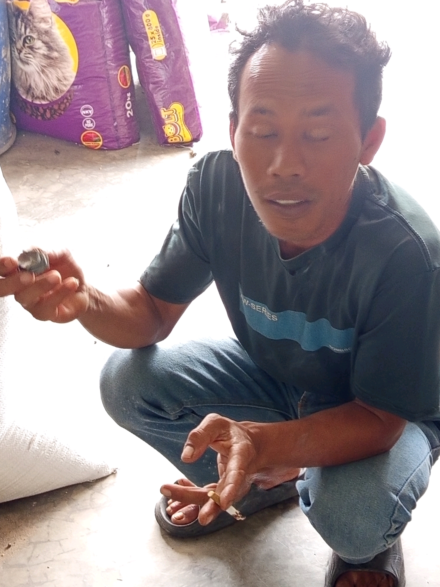 A man is crouching next to several large bags of cat food. He is wearing a dark t-shirt, blue jeans, and black sandals. His eyes are closed, and he appears to be holding a cigarette between his fingers. The bags of cat food are purple and appear to be made of plastic. One bag has a picture of a cat's face on it, and text that reads "20kg." The other bag has text that reads "25 x 300 g inside." The ground is made of concrete. The scene appears to be an outdoor market or storage area in Pulung Kencana, Indonesia.