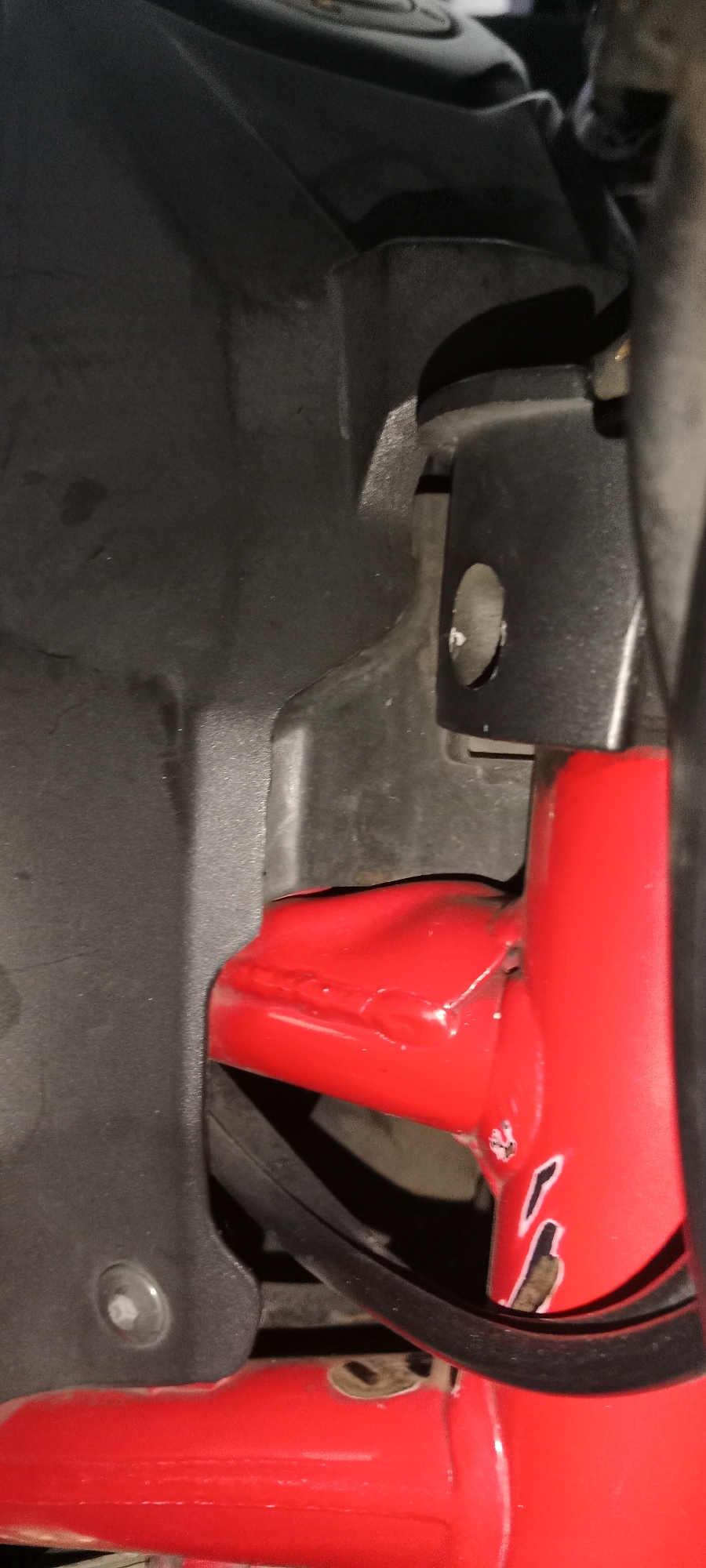 The image shows a close-up, low-angle view of red metal framing, likely part of a vehicle or machinery. A black plastic component is visible on the left, attached by a hex bolt. A black rubber or plastic hose or cable is routed over the red framing. The red framing appears to be painted and shows some signs of wear or damage, with chipped paint revealing a darker material underneath. The overall scene is dimly lit, suggesting an indoor or shaded environment. The exact location within Subang, Indonesia cannot be determined from the visual information provided.