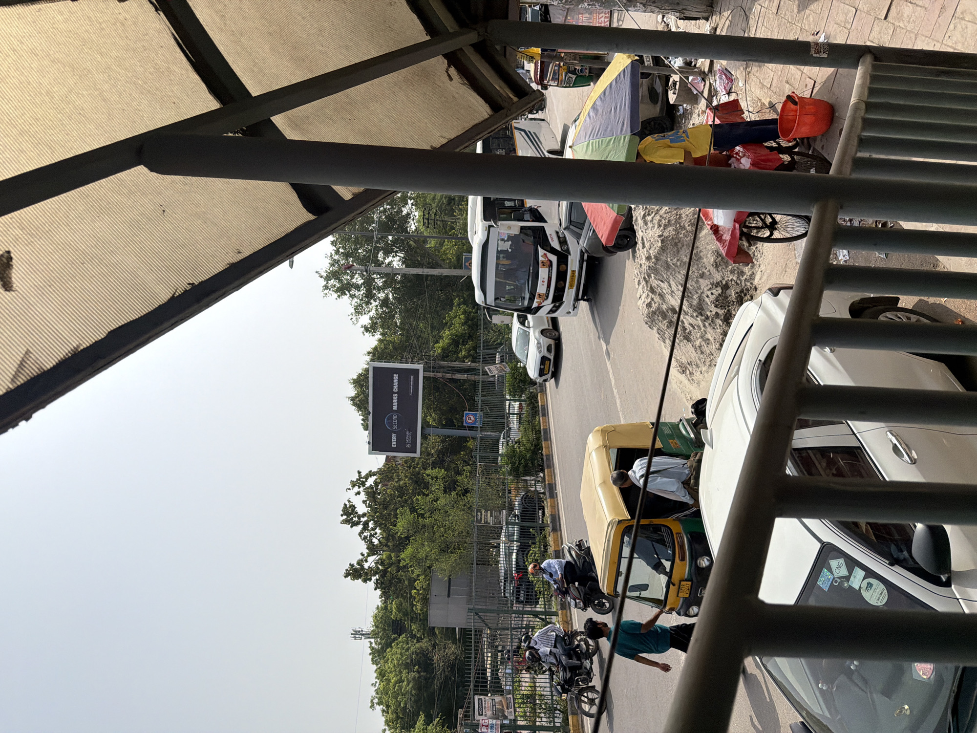 The image depicts a street scene in Delhi, India, likely during daylight hours. In the foreground, a white car is visible through a metal railing. The middle ground shows a busy street with various vehicles, including a yellow and green auto-rickshaw, a white bus, a white sedan, and motorcycles. Several individuals are present, some riding motorcycles, others walking, and one appears to be operating an auto-rickshaw. A street vendor with a cart selling what appears to be ice is also visible. In the background, trees and buildings line the street, and a large billboard with text and graphics is mounted on a pole. The overall environment suggests an urban setting with moderate traffic and pedestrian activity.