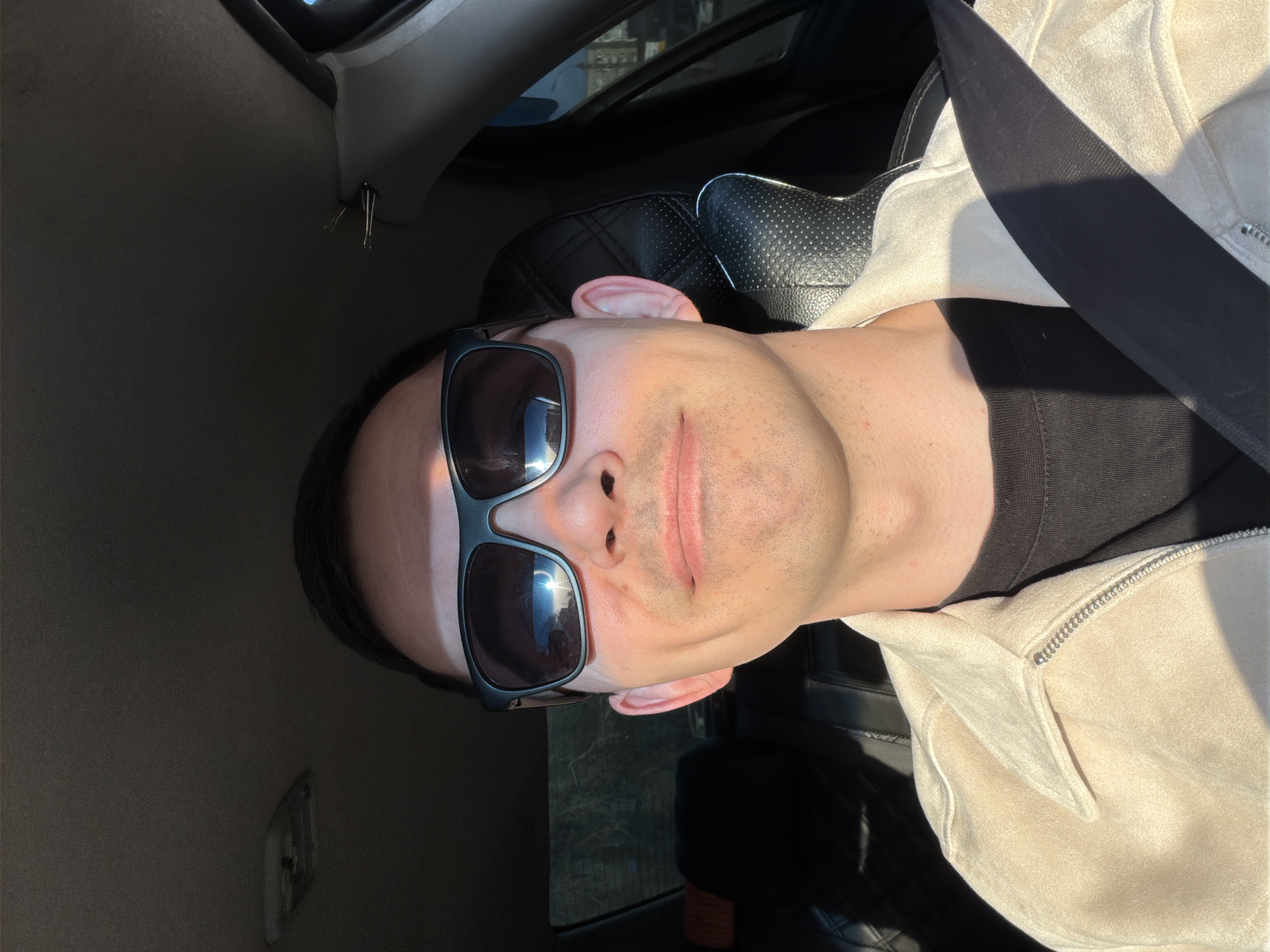 The image is a selfie of a man wearing sunglasses inside a car. He is wearing a black T-shirt and a light-colored jacket or coat, with a seatbelt across his chest. The interior of the car is visible, including the dashboard, a black leather seat, and part of the car window. The light suggests it is daytime. The location in the city of Izhevsk, Russia cannot be determined from the image alone.