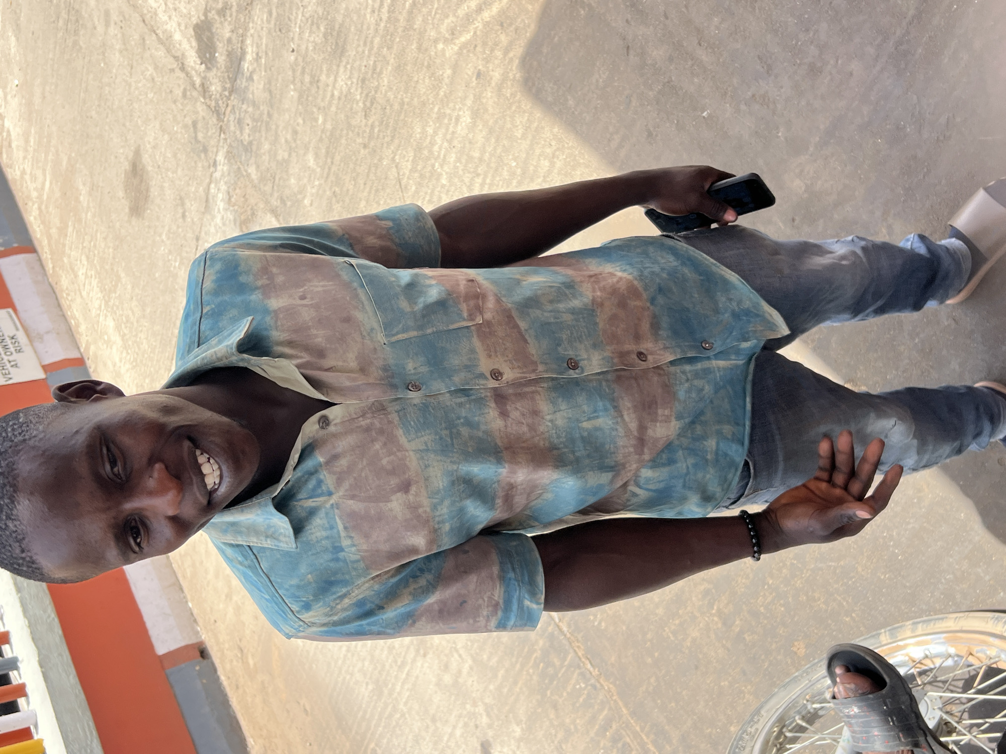 A man stands outdoors in Jos, Nigeria, holding a black smartphone in his left hand. He is wearing a short-sleeved button-down shirt with a striped pattern of light blue and mauve, blue jeans, and sandals. He is smiling and looking at the camera. The ground is paved. A motorcycle wheel is visible near his right foot. A sign is also visible in the periphery.