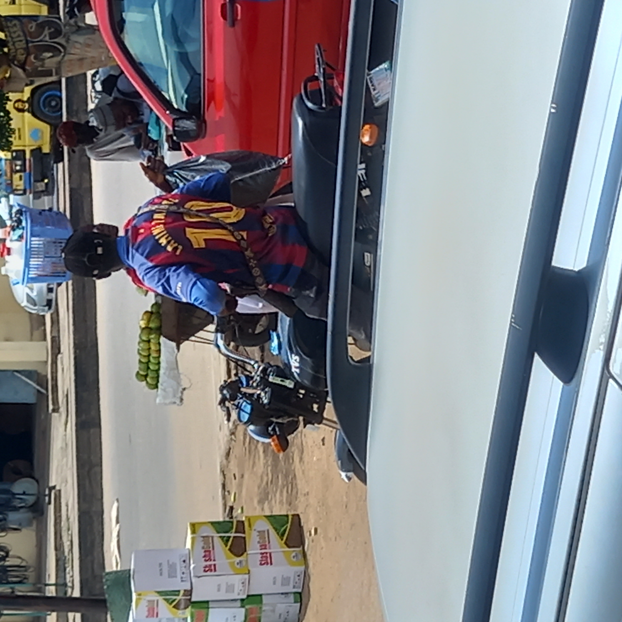 This image captures a vibrant street scene in Dadi Riba, Nigeria, under bright daylight. The central focus is a person, seen from behind, wearing a blue and red striped football jersey with the number "10" and text that appears to be "LM10" on the back, along with a black cap. This individual is seated on a black scooter or motorcycle that serves as a mobile vending station. A visible string of green citrus fruits, likely limes or small lemons, hangs from the rear of the scooter, and a light blue plastic basket is attached to the front.
In the background, a red sedan is parked or moving on the road. Further left, a yellow utility vehicle or bus is visible, displaying partial text including "CRISES" on its side. Several other people are present in the mid-ground, engaging in various activities typical of a bustling market environment. One person near the yellow vehicle is wearing a light-colored garment and a red head covering.
In the lower-left corner (when viewed upright), several stacked cardboard boxes are visible, featuring the text "Say Good" or "Say Gold" on their sides. The right side of the frame is dominated by a large white or light-colored vehicle, possibly a van or bus, partially obscuring the background elements. The scene conveys a sense of everyday commerce and activity on a sunny day in a local street market setting.