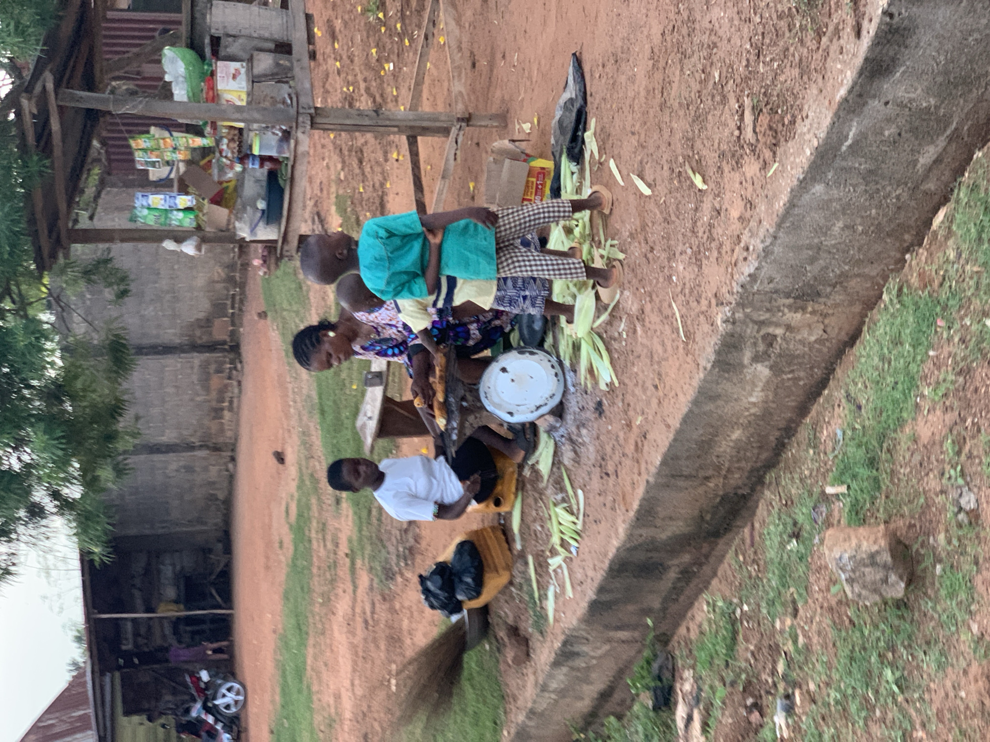 The image depicts an outdoor scene in Gbeleko, Nigeria, likely a market setting. Several people are present, including at least three children and two adults. One child wears a bright teal top, another a yellow shirt, and a third a patterned dress. An adult woman is dressed in a colorful, patterned outfit. Another adult, a young man, wears a white shirt and is seated on a yellow container. He is next to a black trash bag. In the background, a rustic stall is visible, laden with various packaged goods, suggesting it is a small shop. The ground is mostly dirt, with some patches of grass and scattered leaves. A concrete structure runs along the right side of the image, possibly a drainage channel. The overall impression is of a busy, everyday scene. The lighting suggests it is daytime, and the weather appears to be clear. The focus of the activity seems to be around the stall and the people gathered near it, some of whom appear to be interacting with the goods.