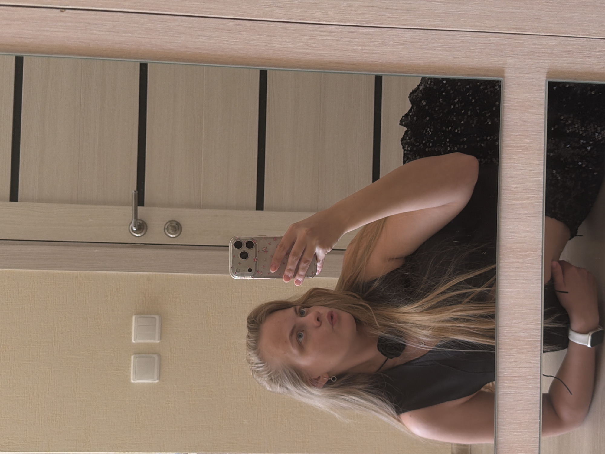 A person with blonde hair and blue eyes is taking a selfie in front of a mirrored closet. The person is wearing a black dress with sequins. The mirrored closet is located in a room with light beige walls and a door with horizontal wood grain. A light switch is visible on the wall to the left of the mirror. A door handle and lock are visible on the door in the mirror. The city of Izhevsk, Russia, cannot be determined from the image.