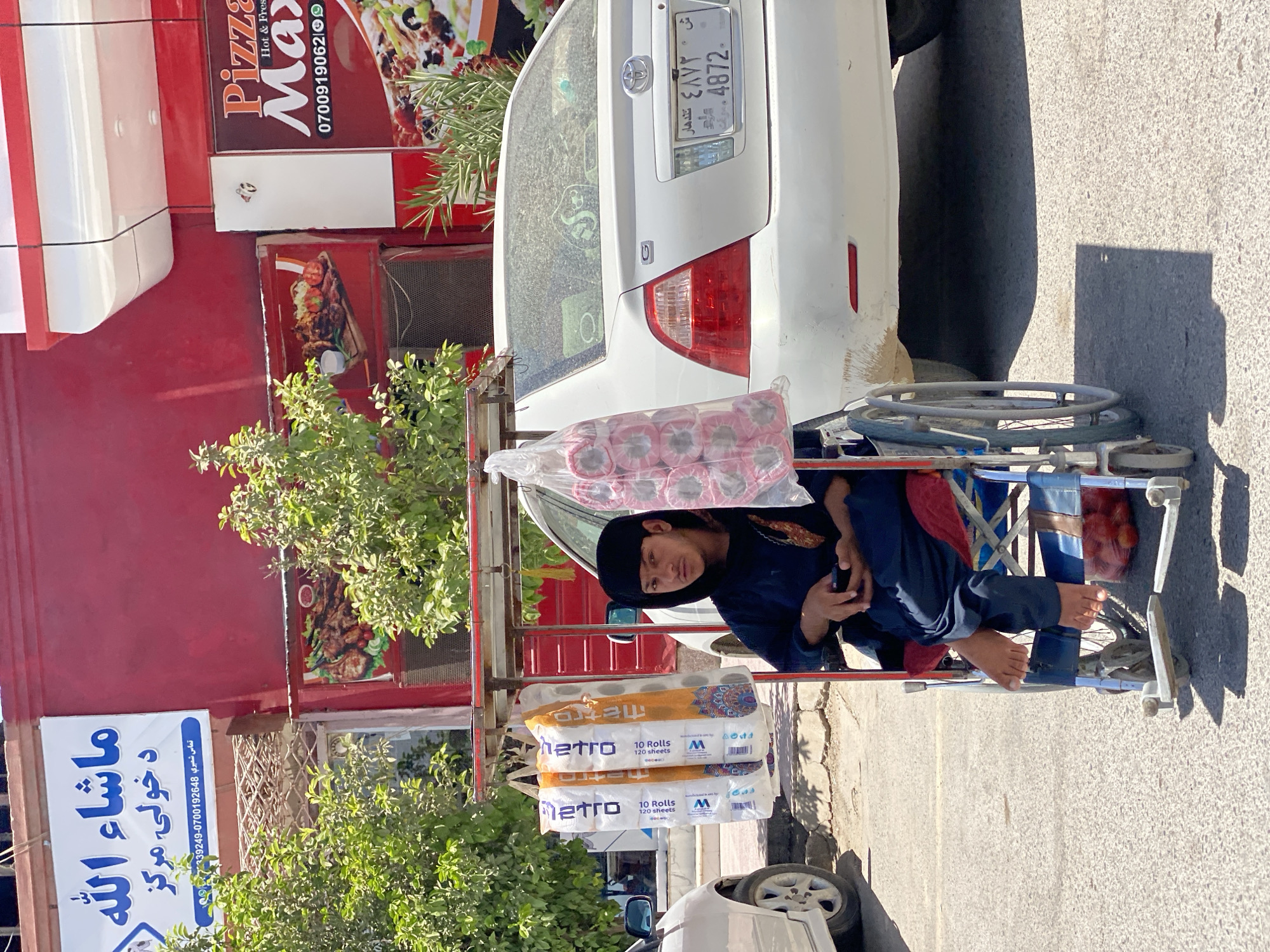 The image depicts a street scene in Kandahar, Afghanistan. In the foreground, a young person is seated in a blue wheelchair, holding a mobile phone. The wheelchair has a red cushioned seat and backrest, and the person is wearing a dark blue traditional outfit and a black head covering. To the left of the wheelchair, a vendor's cart displays several plastic-wrapped rolls of toilet paper. In the background, a white Toyota sedan is parked. Above the sedan, a building with red and white signage is visible. One sign has Arabic script, and another advertises "Pizza Max". The scene is illuminated by bright sunlight, casting shadows on the asphalt road.
