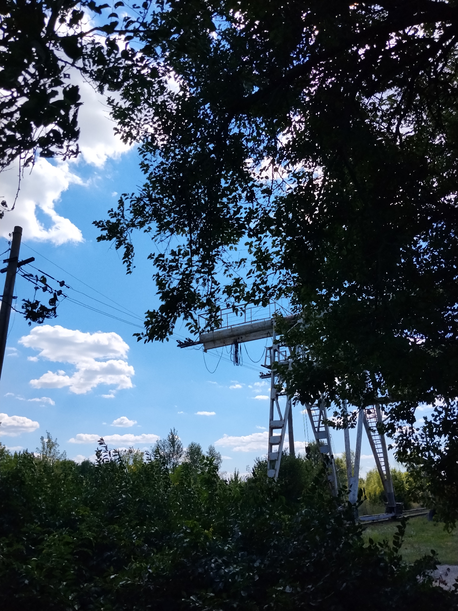 The image, taken in Hoholeve, Ukraine, depicts a bright, partly cloudy blue sky with the sun partially obscured by the dark silhouettes of tree branches and foliage in the foreground and midground. A utility pole with power lines runs diagonally from the left side of the frame upwards, connecting to unseen structures. In the background, a large industrial structure, possibly a crane or gantry, stands prominently with its metal framework and horizontal beam visible. This structure is situated amidst a backdrop of green trees and vegetation. The scene appears to be an outdoor, possibly rural or industrial, setting under daylight conditions.