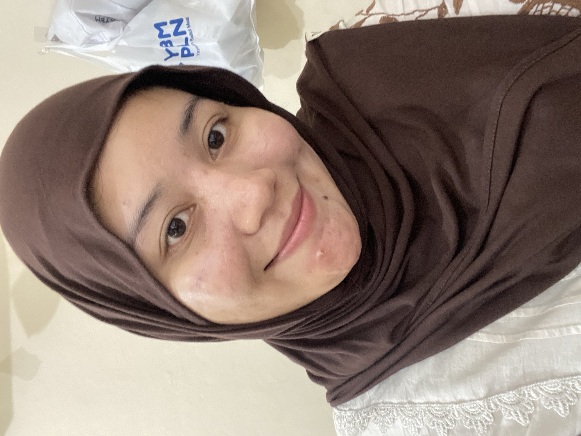 This is a medium-close up, selfie-style photograph of a young Indonesian woman wearing a brown hijab and a white lacy top. She is looking at the camera with a slight smile, and her face is illuminated by soft lighting. The background appears to be an indoor wall. There are no distinctive landmarks in the image that would allow for precise geolocation to Banjarbaru, Indonesia.