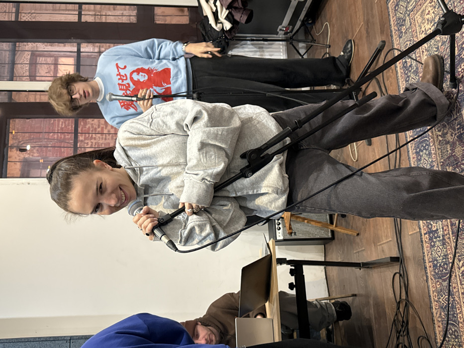 The image depicts an indoor scene in Moscow, Russia, likely a studio, rehearsal space, or informal performance venue.
The central figure is a young adult female, light skin tone, with dark hair tied in a high ponytail. She is smiling broadly and looking towards the viewer, holding a silver-colored microphone in her right hand near her mouth. She wears an oversized light grey hooded sweatshirt, dark grey wide-leg jeans cuffed at the ankles, and brown work boots. Several rings adorn her fingers. A black microphone stand is positioned in front of her.
Behind and slightly to the right of the central figure stands a young adult male, light skin tone, wearing glasses. He has dark hair and a neutral expression, holding a microphone attached to another black stand. He is dressed in a light blue long-sleeved sweatshirt featuring a red graphic of a woman's face and Japanese text (愛見せて, "Show me love"). He wears dark trousers and dark shoes.
To the far left, a person in a blue hooded sweatshirt is partially visible. Further left, seated at a light wooden table with black metal legs, is a young adult male, light skin tone, wearing a brown hooded sweatshirt and a dark beanie. He is focused on a silver laptop. Another black laptop is also present on the table.
The room features white walls on the left and large windows on the right, through which an exterior brick building is visible. The floor consists of dark wooden planks, partially covered by an ornate, multi-colored patterned rug. Equipment visible includes a small silver amplifier with control knobs on the floor near the table, and a black road case or equipment box behind the second male figure. Numerous black cables are distributed across the floor, connecting various pieces of equipment. The overall environment suggests an audio recording or live music setup.