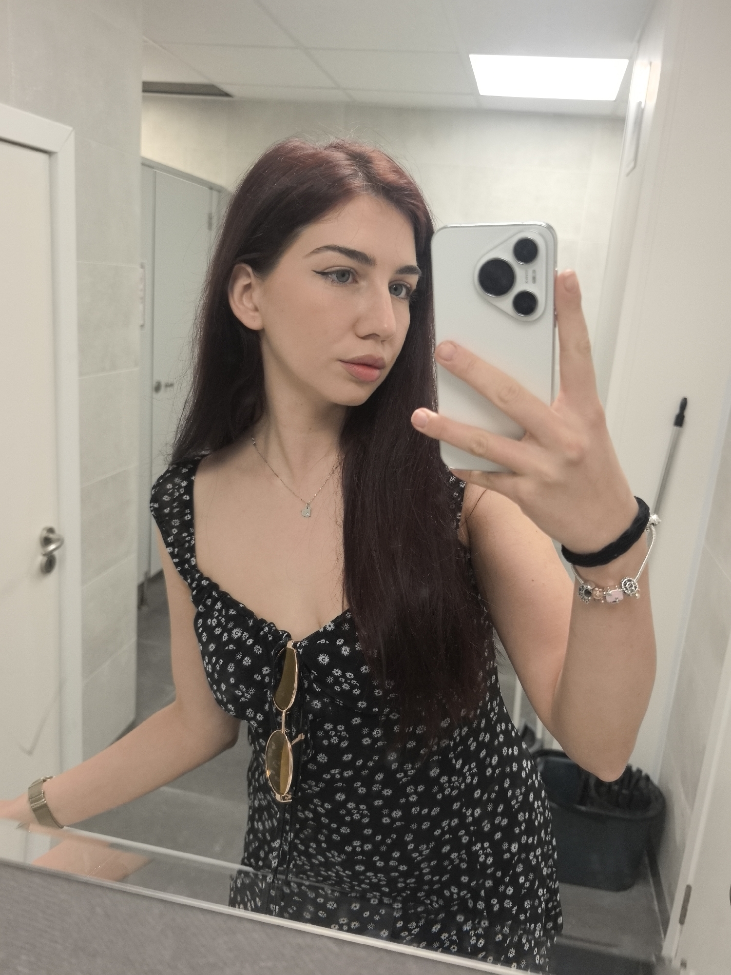 A young woman with long, dark brown hair and light eyes is captured in a mirror selfie within what appears to be a public restroom or changing room in Bogomilovo, Bulgaria. She is wearing a black, sleeveless dress with a small white floral pattern. A pair of gold-rimmed sunglasses hangs from the neckline of her dress.
Her left hand holds a silver smartphone, obscuring part of her face, while her right arm, adorned with a silver charm bracelet and a black hair tie/bracelet, rests casually. A watch with a metallic band is visible on her right wrist, closer to the mirror's edge. A delicate silver necklace with a small heart pendant is visible against her chest.
The background shows light-colored tiled walls and two white doors, resembling restroom stalls, reflected in the mirror. A rectangular ceiling light fixture illuminates the space. In the reflection behind her, a dark bin or bucket and a white stick (possibly a mop handle) are visible, reinforcing the utilitarian setting. The woman's expression is neutral and composed, with her gaze directed slightly away from the camera. There is no visible text in the image.