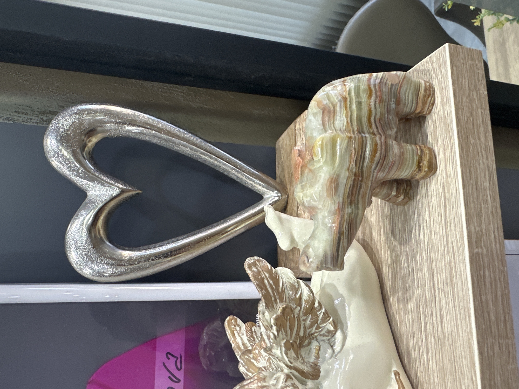 A collection of decorative objects is displayed on a light-colored wooden shelf. Dominating the background is a large, silver, hollow heart sculpture with a textured surface. To the left of the heart is a creamy white sculpture of angel wings. In front of the wings, and slightly to the right, is a small sculpture of an animal, possibly a bear or a pig, carved from layered, multi-colored stone, exhibiting bands of white, green, orange, and brown. The shelf itself has a visible wood grain pattern and appears to be part of a larger display unit. The background behind the objects is dark, with vertical white blinds visible on the far right. A portion of a dark framed structure is also present to the right of the heart sculpture. The image does not contain any information indicating its location in Haskovo, Bulgaria.