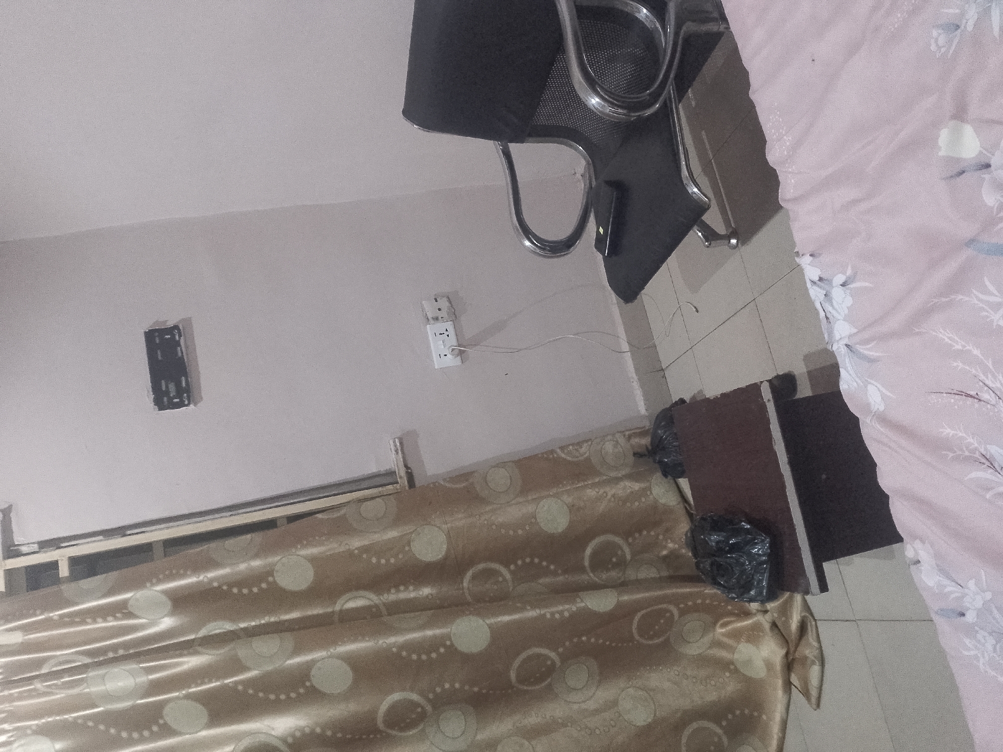 The image depicts an indoor scene, possibly a bedroom or living area. A black office chair with a metal frame is partially visible in the upper portion of the frame. On a light-colored wall, a white electrical outlet with a cord plugged in is mounted. To the left of the outlet, a black wall mount for a television is visible. In the lower part of the image, a beige curtain with a circular pattern is drawn, covering a window or alcove. Next to the curtain, a dark brown wooden cabinet with black trash bags placed on it is visible. The floor appears to be tiled. A pink blanket with a floral pattern is draped over a surface on the right side of the image. The overall lighting suggests an indoor environment.