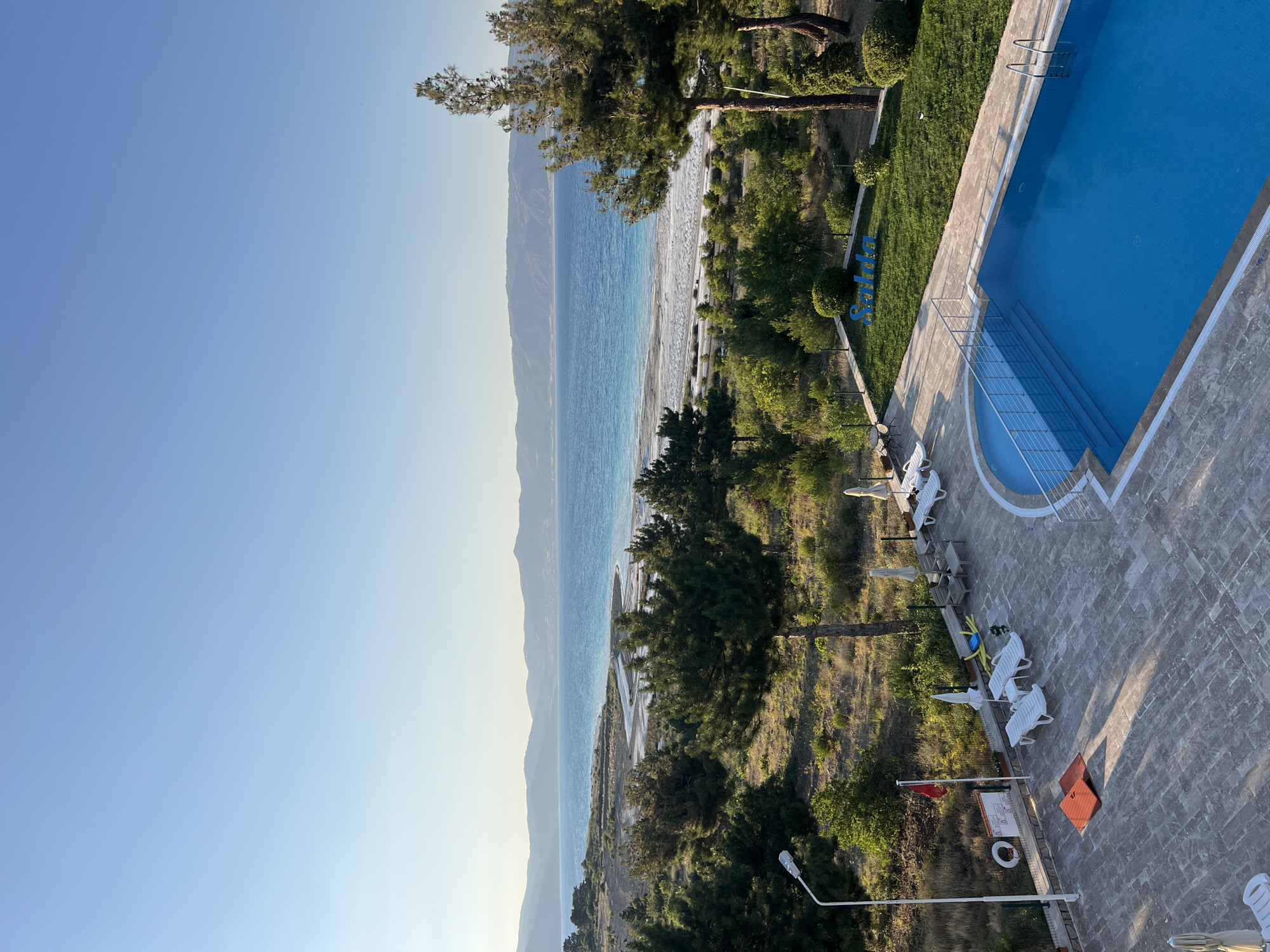This high-angle outdoor shot captures a serene scene at Salda Lake in Yeşilova, Turkey. In the foreground, a brightly lit blue swimming pool with metal railings and steps sits on a stone-tiled patio, accompanied by several white lounge chairs and umbrellas. Further back, lush green trees and vegetation lead to the sandy shores of Salda Lake, a body of water with distinct turquoise and blue hues. The lake is bordered by a white, sandy beach and a distant mountain range under a clear, pale blue sky. A sign with the word "Salda" in blue lettering is visible on a manicured lawn. A flagpole with a red flag and a life preserver ring are also present near the patio.