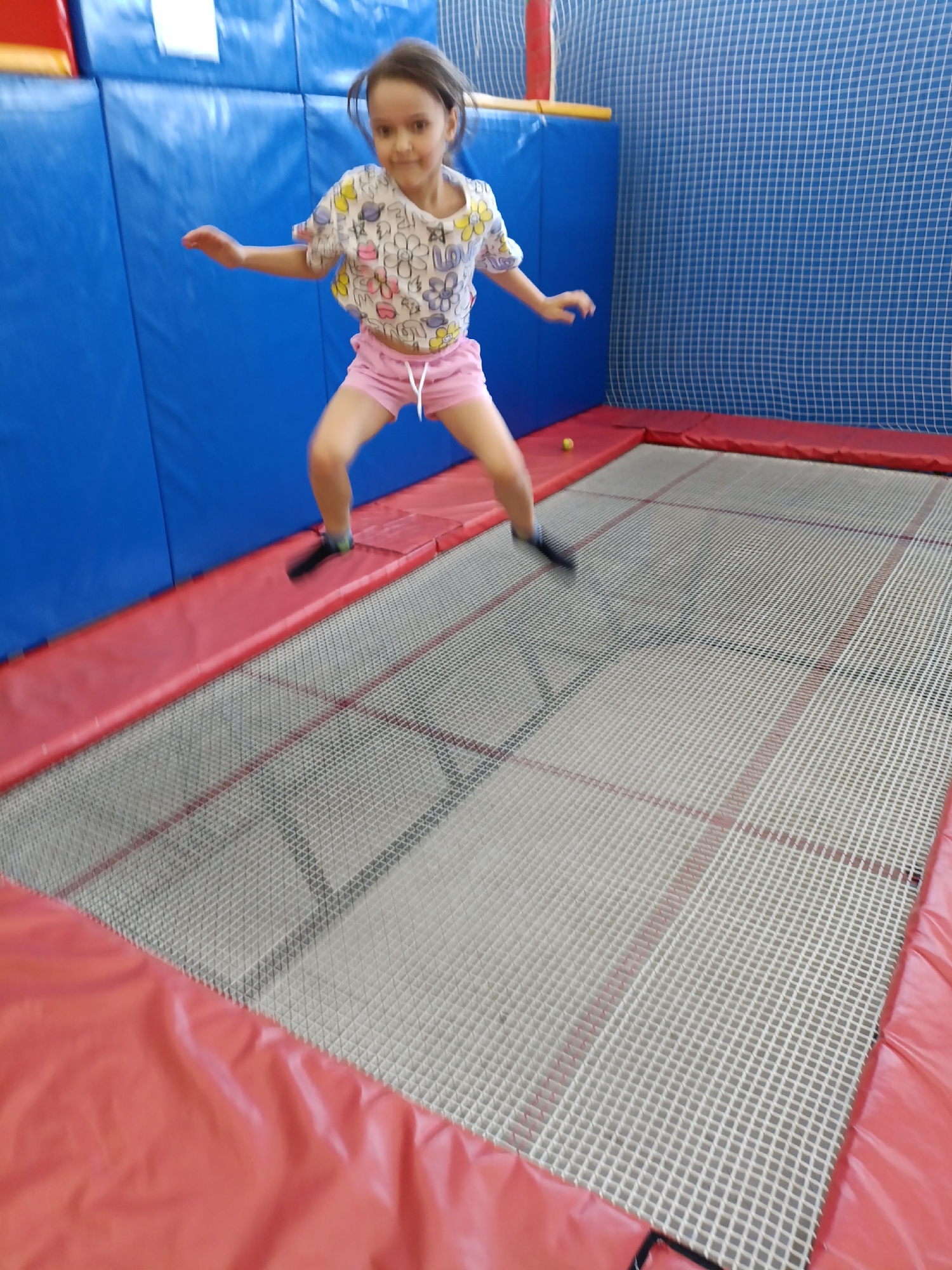 A young girl is jumping on a trampoline. She is wearing a white t-shirt with colorful prints and pink shorts. The trampoline has a red safety pad around the edge and a netting surface. The background features blue padded walls and a blue netting. The location is identified as the city of Chaikovsky, Russia.