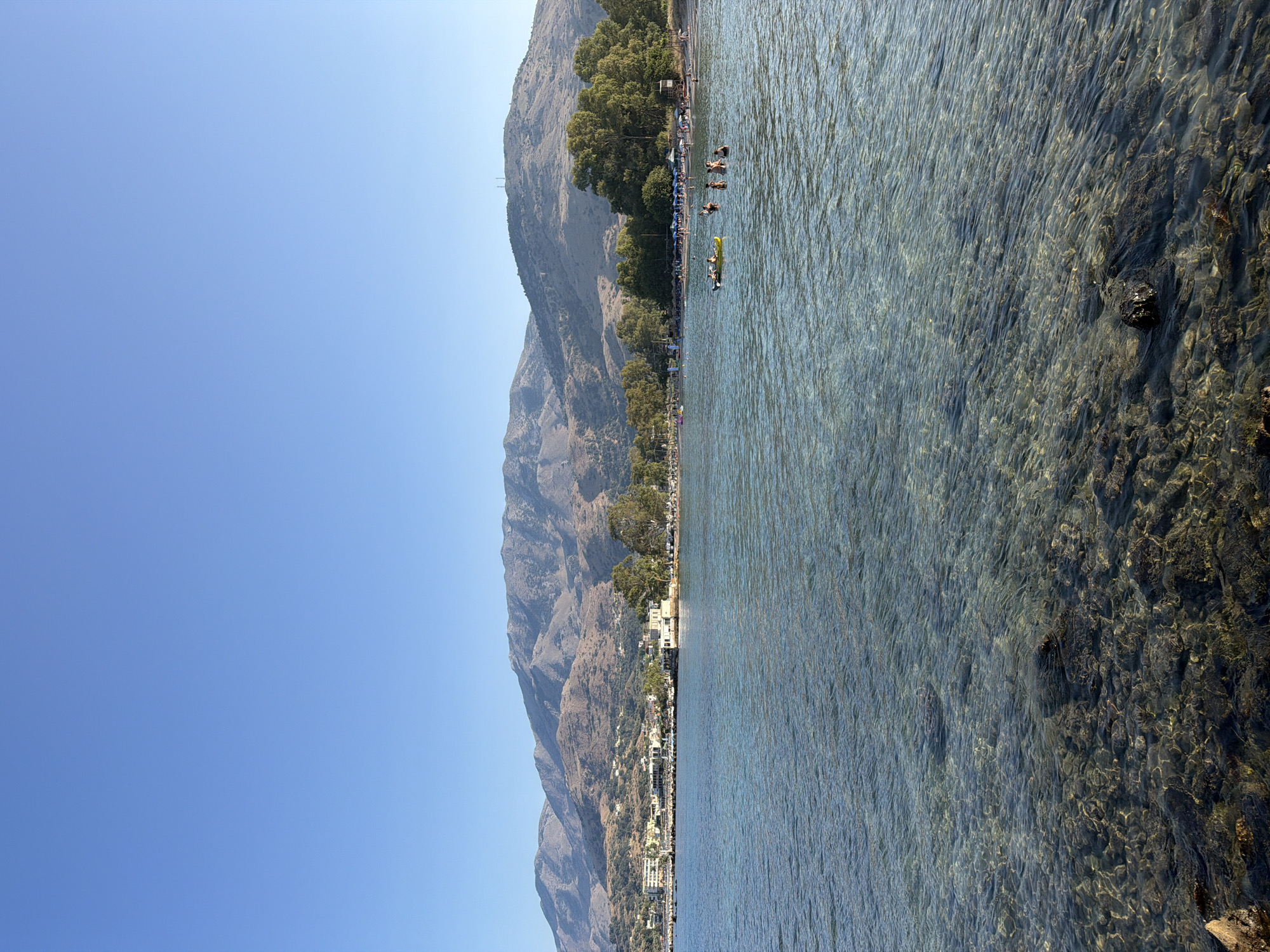This image depicts a sunny day at a beach in Georgioupoli, Greece. In the foreground, the clear, shallow water reveals a rocky seabed. In the mid-ground, several people are visible in the water, some swimming and others on a yellow inflatable. Further back, a sandy beach is lined with umbrellas and trees, and beyond that, buildings of a town are nestled against a backdrop of large, dry, mountainous terrain. The sky is a clear, bright blue.