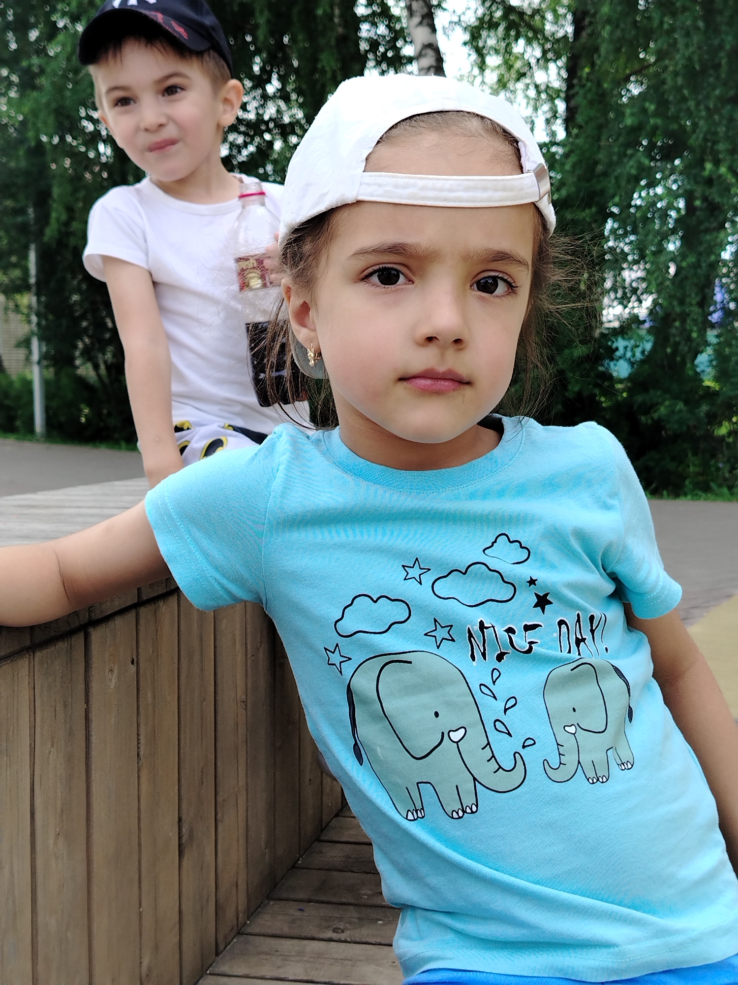In the city of Yanaul, Russia, two children are pictured outdoors. A young girl in the foreground, wearing a light blue t-shirt with a graphic of elephants and clouds, and a white baseball cap, looks directly at the camera. Behind her, a boy in a white t-shirt and a black baseball cap is partially visible, looking to his left. They appear to be near a wooden structure, possibly a bench or playground equipment, with trees and foliage in the background.