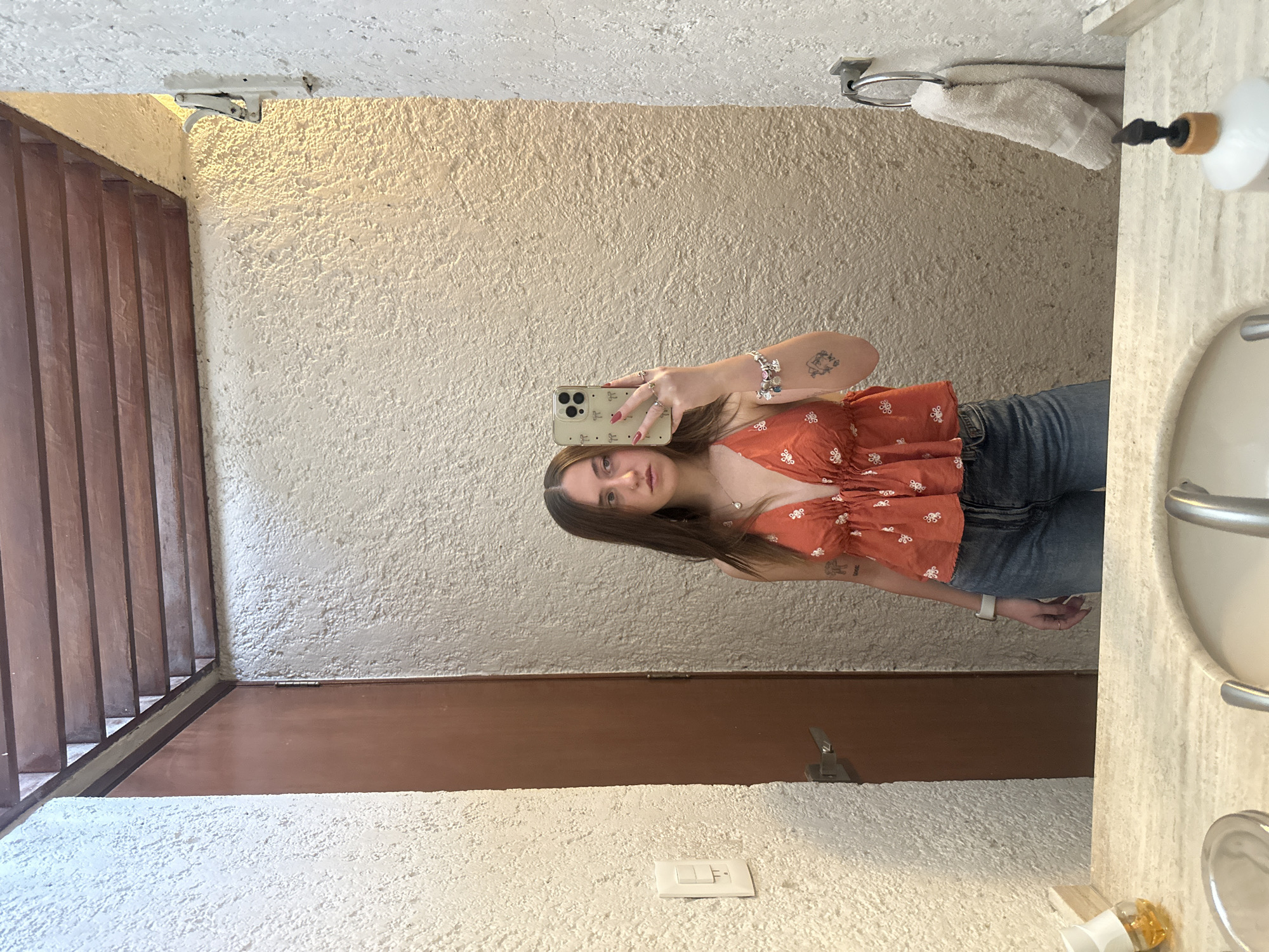 A young woman is taking a selfie in a bathroom in Naucalpan de Juárez, Mexico. She is wearing an orange floral top and jeans. The bathroom has a light-colored, textured wall and a brown door. There is a sink to the right of the woman, and a towel rack above the sink. A light switch can be seen on the wall near the door. There are wooden slats visible on the left side of the frame.