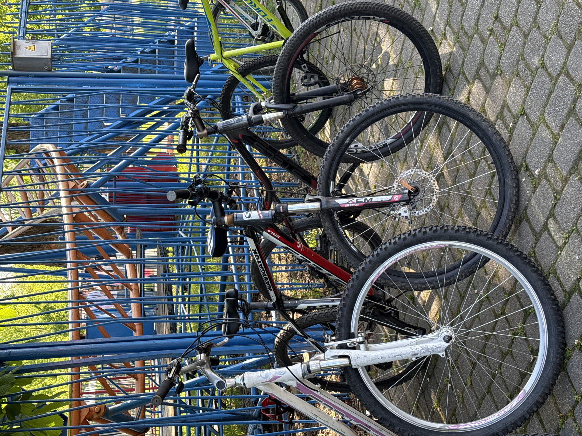 The image shows a row of bicycles parked in a blue metal bike rack on a paved surface. There are at least three bicycles, with the front wheels of each bicycle visible. The first bike is green, the second is black and red, and the third is white and purple. The bike rack is constructed from blue metal bars, and behind it, there is a taller orange and blue metal structure resembling stairs. The paved surface is made of interlocking concrete blocks. The location is Unknown, Unknown.