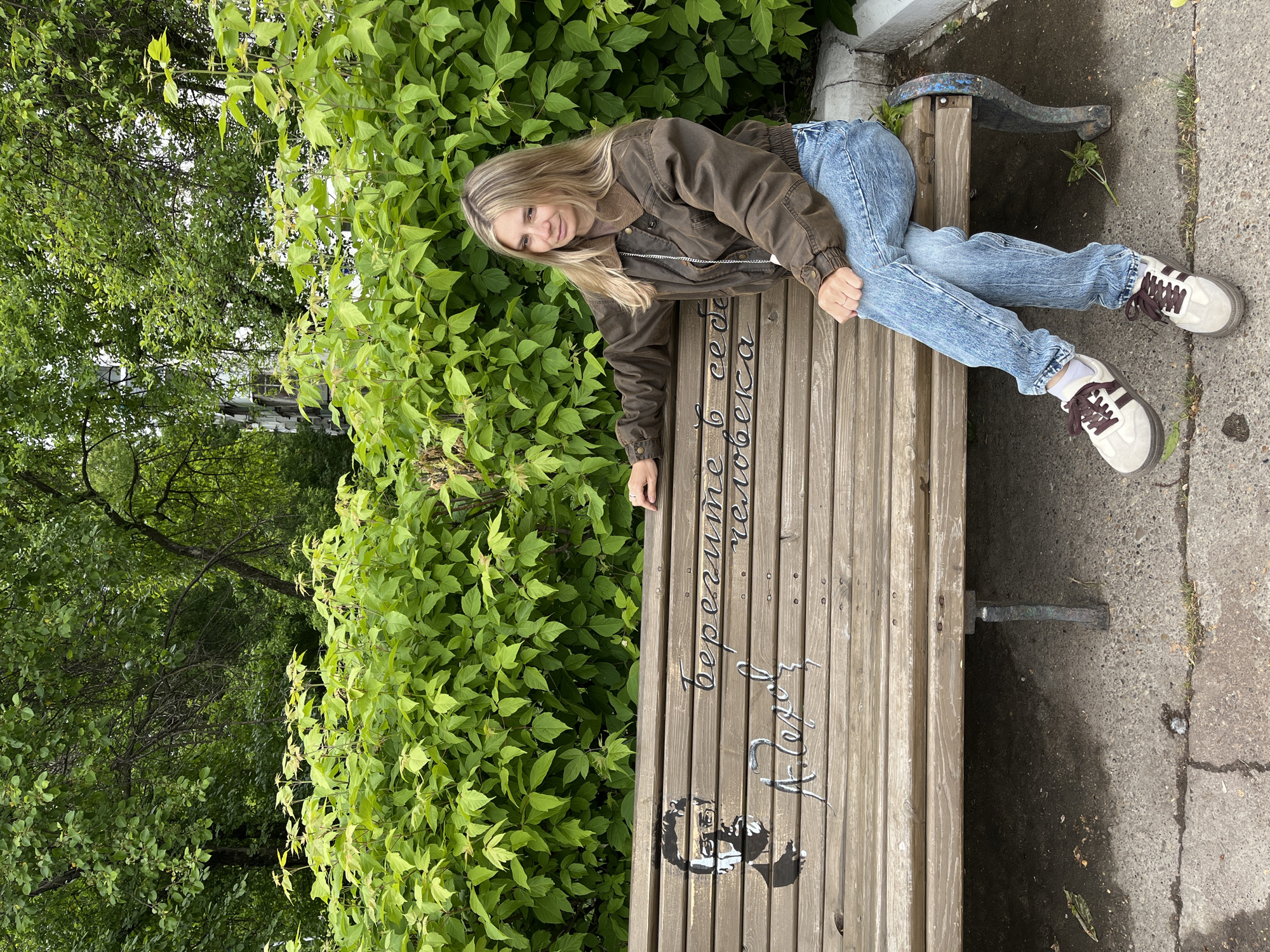 A woman is lying on a bench in Chaikovsky, Russia. The bench is made of wood and has graffiti on it. The woman is wearing a brown jacket, blue jeans, and sneakers. She is lying on her side, looking at the camera. Behind her is a wall of green leaves. To the left, trees can be seen. To the right of the bench is a concrete slab.