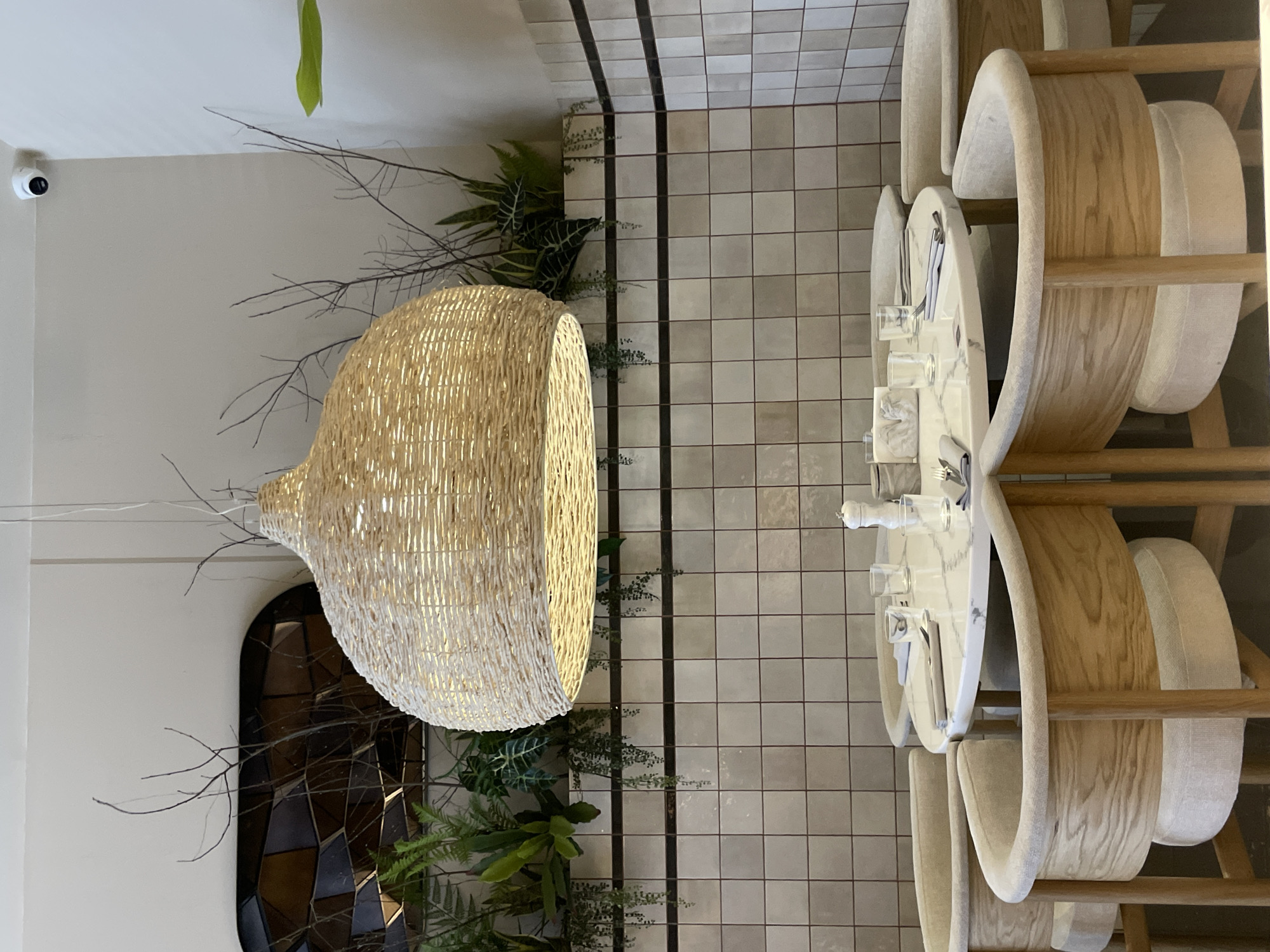 An interior shot shows a dining area. A large, woven, teardrop-shaped lamp hangs from the ceiling, near a dark archway surrounded by plant decorations. The floor is tiled in a grid pattern. To the right, a table is set with silverware and glasses, surrounded by chairs with light-colored wooden frames and padded seats. A security camera is mounted in the corner. The location and specific city are not ascertainable from the image.
