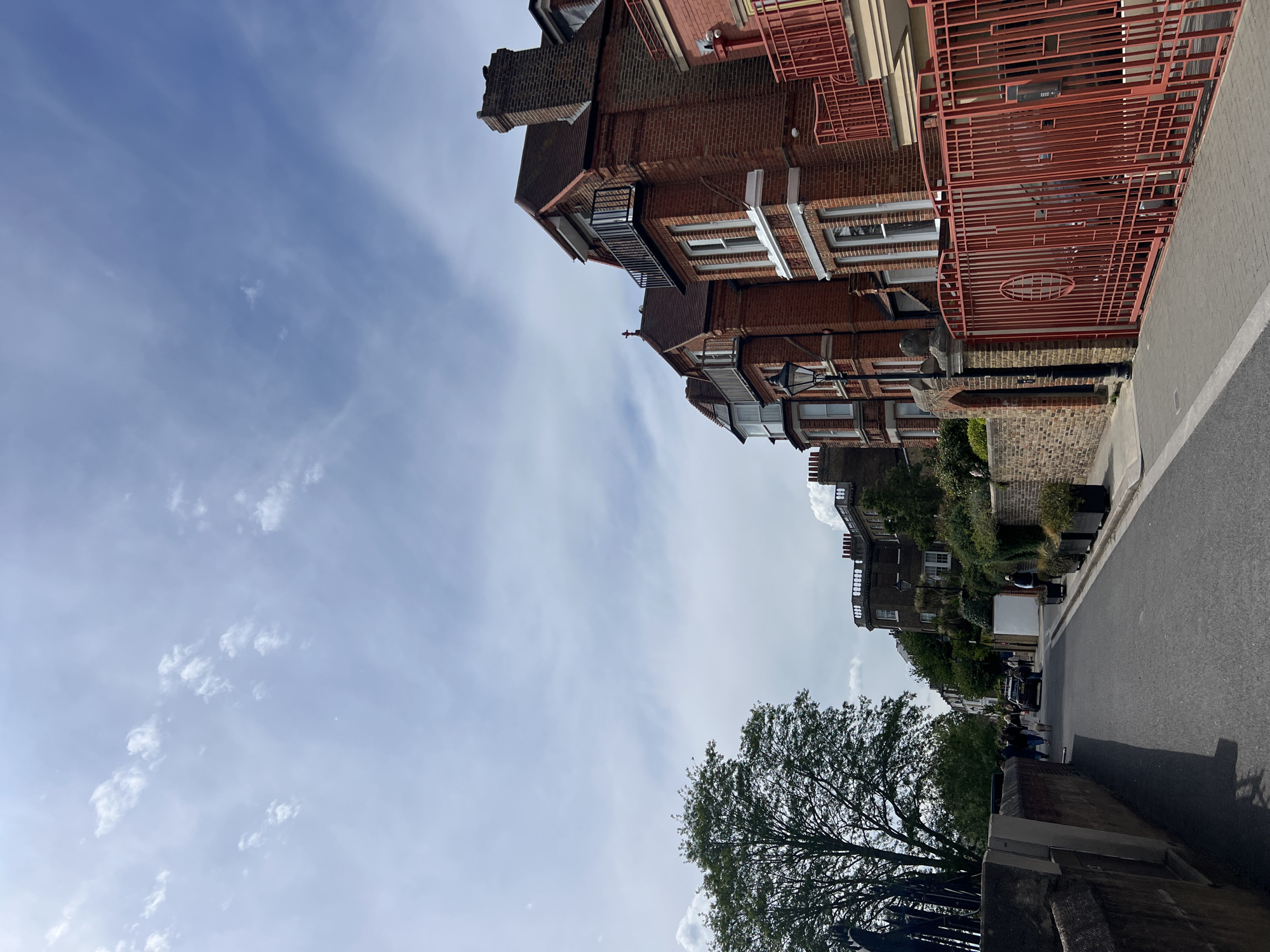 The image shows a street scene in London, United Kingdom. The view is angled upwards towards a partly cloudy sky. On the right, a residential street with brick buildings featuring architectural details like bay windows, iron railings, and chimneys is visible. The road is paved, and a sidewalk is present. A large tree is on the lower right, partially obscuring the view further down the street. Several people, cars and a white van can be seen further down the street.