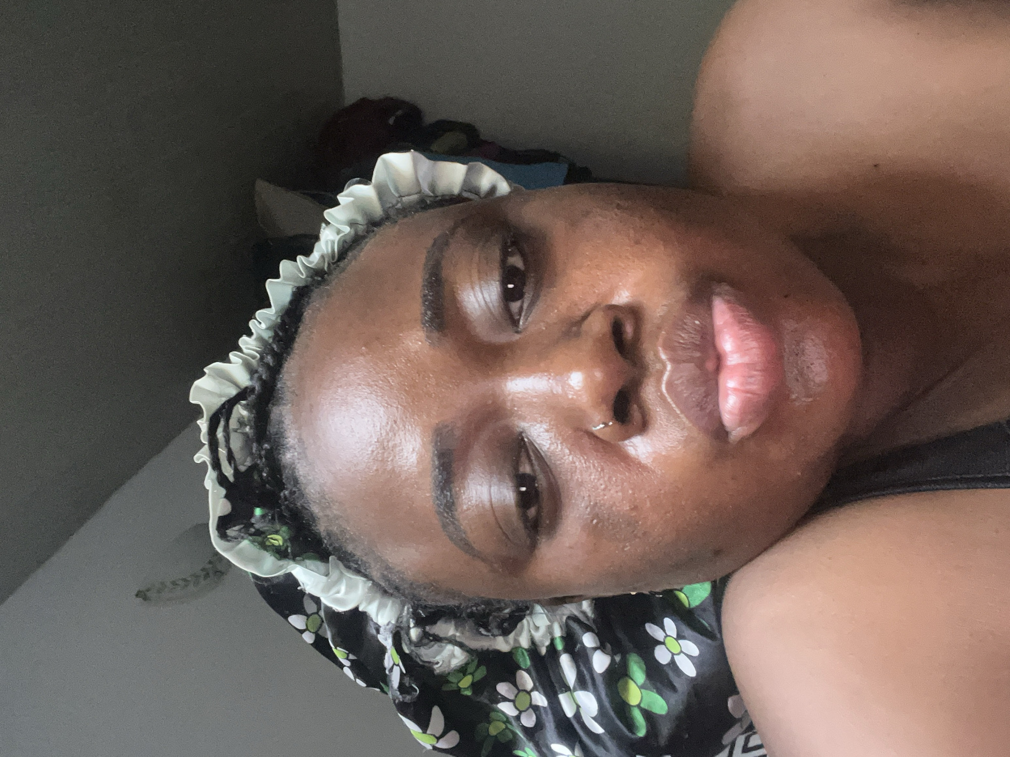 A close-up, angled selfie shows a Black woman with a nose ring wearing a floral bonnet with a ruffled white trim. Her skin appears to have a slight sheen. The background is blurry and indistinct. The image does not provide details to determine a specific location within Abuja, Nigeria.