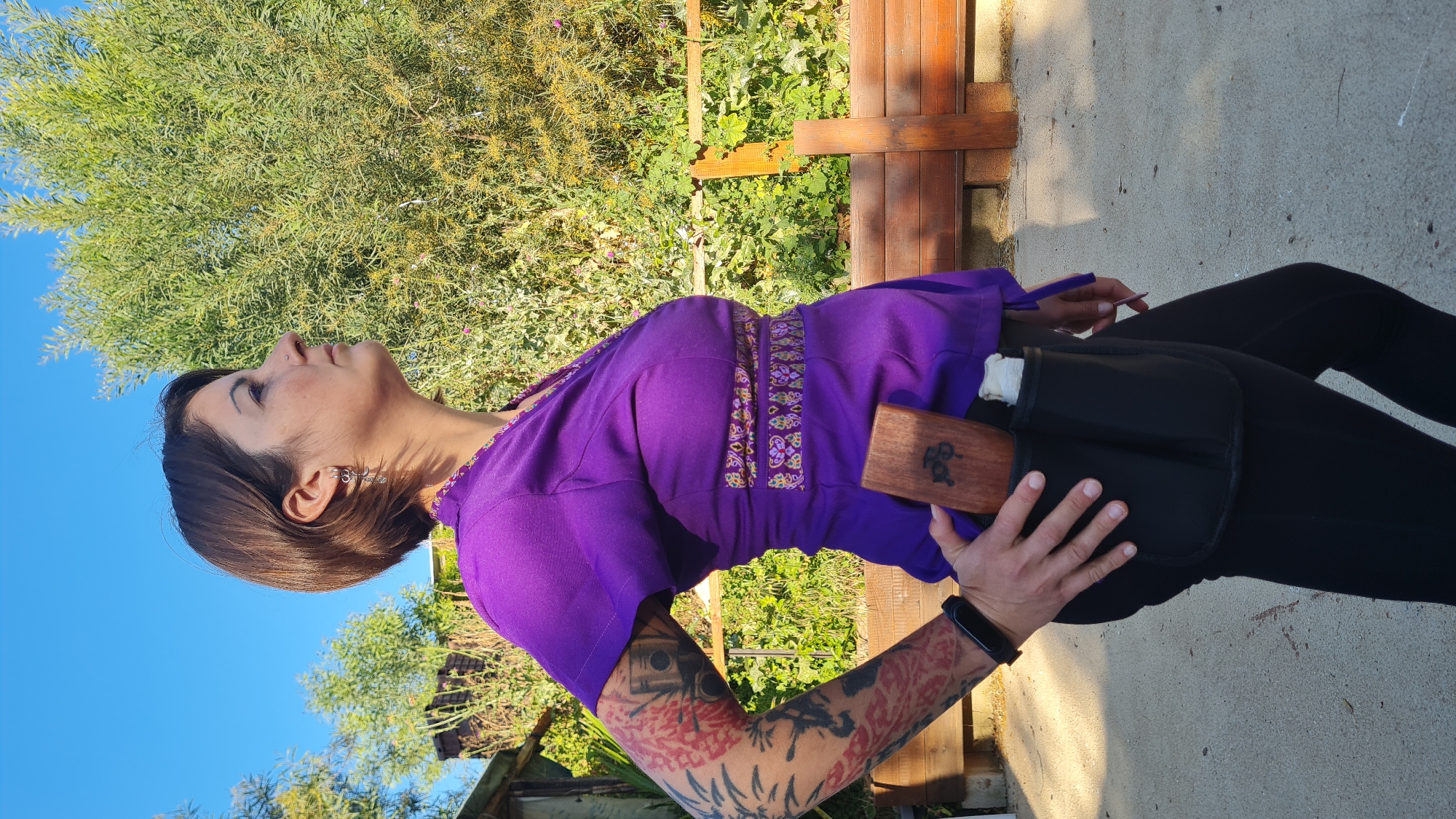 The image captures an adult woman standing outdoors, looking upwards and slightly to her left, under a clear blue sky. She is presented in profile from the torso up, with her left side facing the viewer. Her dark hair is styled short, and she has an earring with a spiraling design in her left ear and a small nose piercing.
She wears a vibrant purple top featuring colorful, intricate embroidered patterns along the neckline and waistline. Her visible left arm is heavily tattooed with diverse, colorful designs, including what appears to be a camera and floral motifs. A dark digital wristband is on her left wrist.
Around her waist, she has a black utility belt or pouch system. Her right hand rests on a rectangular wooden block that is integrated into this belt and features stylized carvings or symbols on its surface.
The background is dominated by lush green foliage, likely trees or large shrubs, under a bright, cloudless blue sky. A rustic wooden structure, possibly part of a bench or fence, is visible behind her, and the ground appears to be a light-colored concrete surface. The strong, direct sunlight indicates a clear day, likely mid-morning or afternoon, creating visible shadows. The setting suggests a natural or garden environment, consistent with the Lachish Regional Council in Israel. No discernible text is visible.