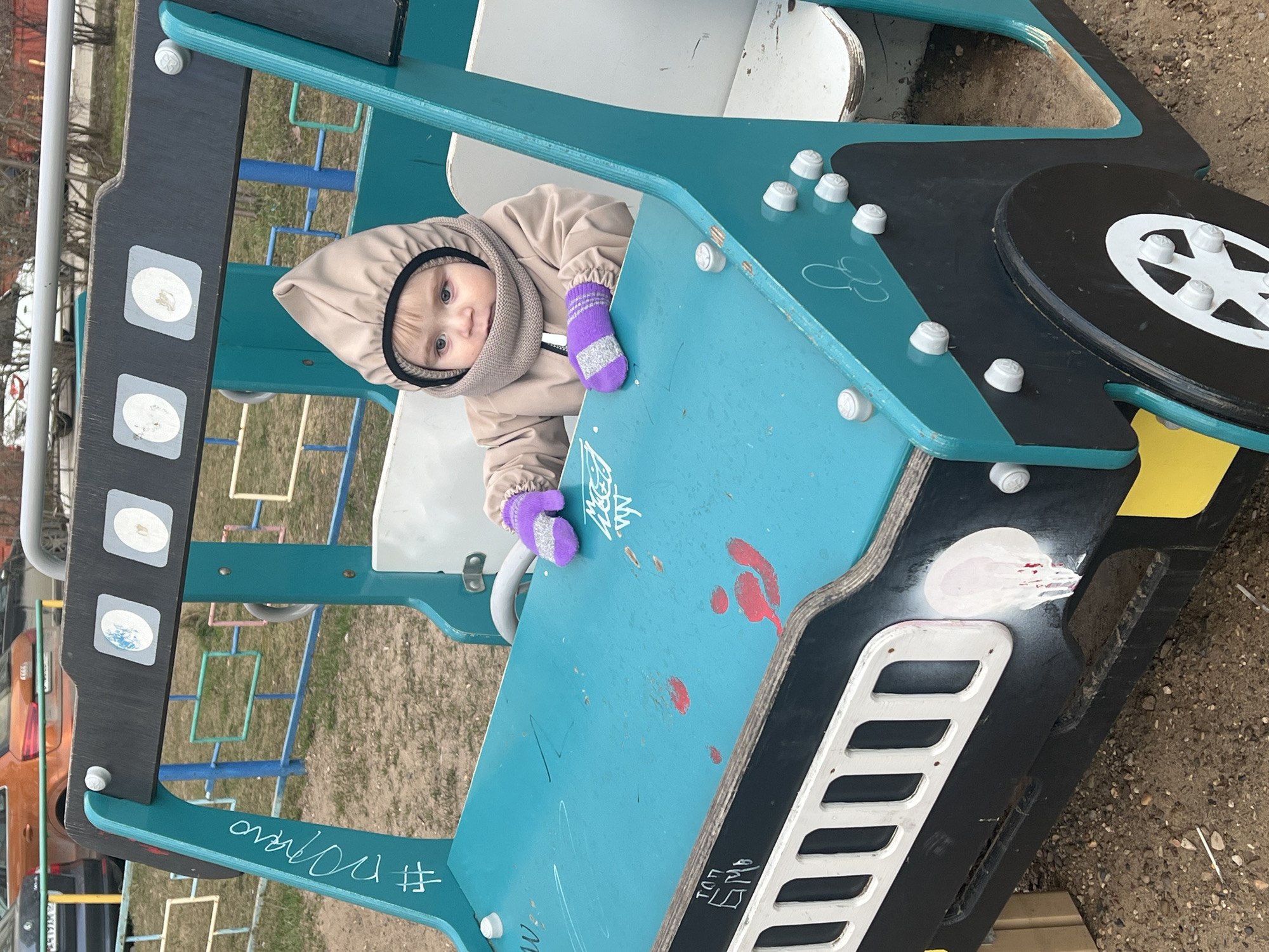 An overhead, slightly angled shot captures a young child in a beige hooded jacket and purple mittens, peeking out from inside a blue and black playground structure designed to resemble a vehicle. The child has light hair and blue eyes, looking directly at the camera with a curious expression.
The playground equipment, situated in an outdoor setting in Perm, Russia, appears worn with various scuffs, scratches, and graffiti. The ground beneath is dirt and gravel. The vehicle-shaped structure features a prominent blue top panel, a black base, a black circle resembling a wheel with white spoke details, and a white slatted grill at the front. Several white, cap-like fixtures are visible on the blue surface, possibly representing controls or decorative elements. Red splatters are present on the blue panel, suggesting old paint or other stains.
In the background, bare trees and other playground components like metal bars and a ladder-like structure are visible. The child's warm attire suggests cool weather, likely early spring or late autumn.
Visible text includes a white hashtag and word, possibly "#ovovo", inscribed on the side of the blue panel. Near the grill, some faint white characters, potentially "ЫЧП" or similar, are discernible.