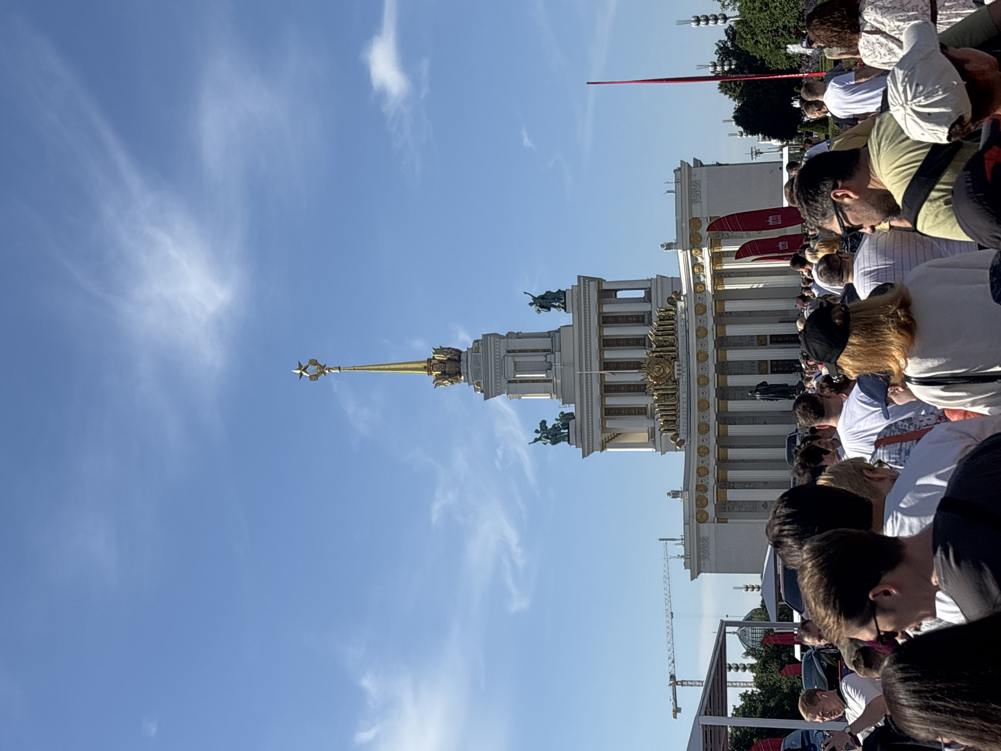 The image captures a sunny day at the VDNKh (Exhibition of Achievements of National Economy) in Moscow, Russia. In the background, the iconic Pavilion No. 57 "Russia - My History" is prominently featured. The grand structure, adorned with golden details and topped with a star, stands against a clear blue sky with wispy white clouds. Statues of figures in dramatic poses are visible on top of the pavilion.
In the foreground, a large crowd of people are gathered, facing the pavilion. Many are looking up at the architecture, while others are engaged in conversation or taking photographs. The diversity of the crowd suggests a public event or a popular tourist attraction. People are dressed in casual summer attire.
To the left of the pavilion, a construction crane is visible, hinting at ongoing development or restoration work at the exhibition grounds. Red flags and banners, some displaying Cyrillic text, are scattered throughout the scene, adding pops of color and indicating a possible event or celebration. Lush green trees frame the sides of the image, providing a natural contrast to the monumental architecture and the bustling crowd. The overall atmosphere appears lively and vibrant.