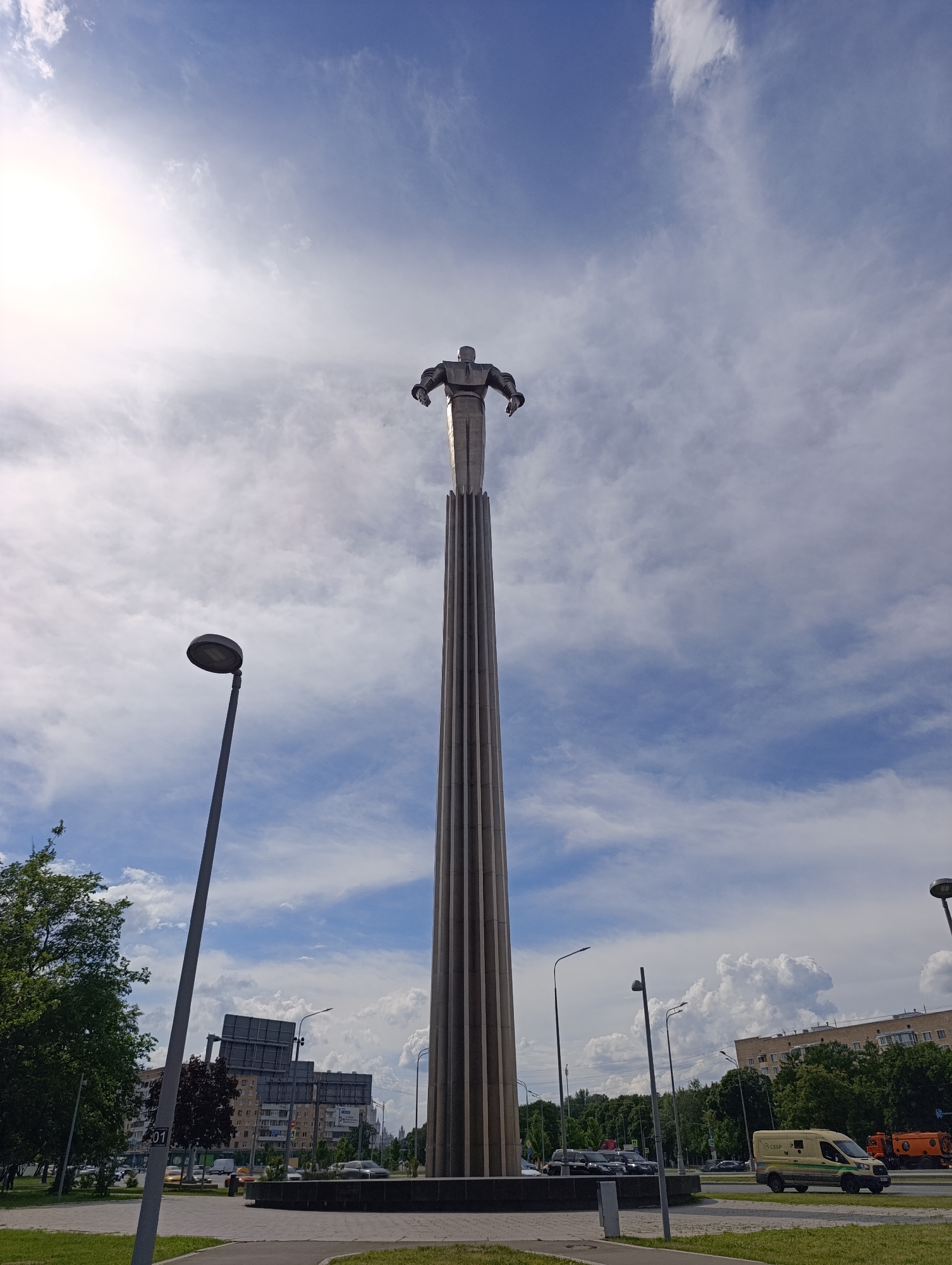 A towering monument, identified as the Monument to the Conquerors of Space (Cosmonauts Monument) in Moscow, Russia, stands prominently against a bright, partly cloudy daytime sky. The monument features a rocket-like pedestal, tall and slender with ribbed detailing, culminating in a statue of a human figure with outstretched arms, seemingly taking flight. The figure appears to be a cosmonaut.
The sun is intensely bright on the left side of the frame, casting a strong glare and illuminating the clouds which are a mix of white cumulus and wispy cirrus against a blue sky.
In the midground, the urban environment of Moscow is visible. A tall, dark grey lamppost stands on the left, with lush green trees and multi-story buildings further behind it. A road with vehicles, including cars and a white van, runs across the scene. Another lamppost is visible near the base of the monument on the right. The white van has green stripes and the Cyrillic text "СБЕР" (SBER) written on its side, identifying it as a Sberbank vehicle. An orange municipal or construction vehicle is also visible on the road to the right. The foreground consists of a paved path and green grass. The scene depicts a sunny, active day in the city, with the monument serving as a focal point of national pride and technological achievement.