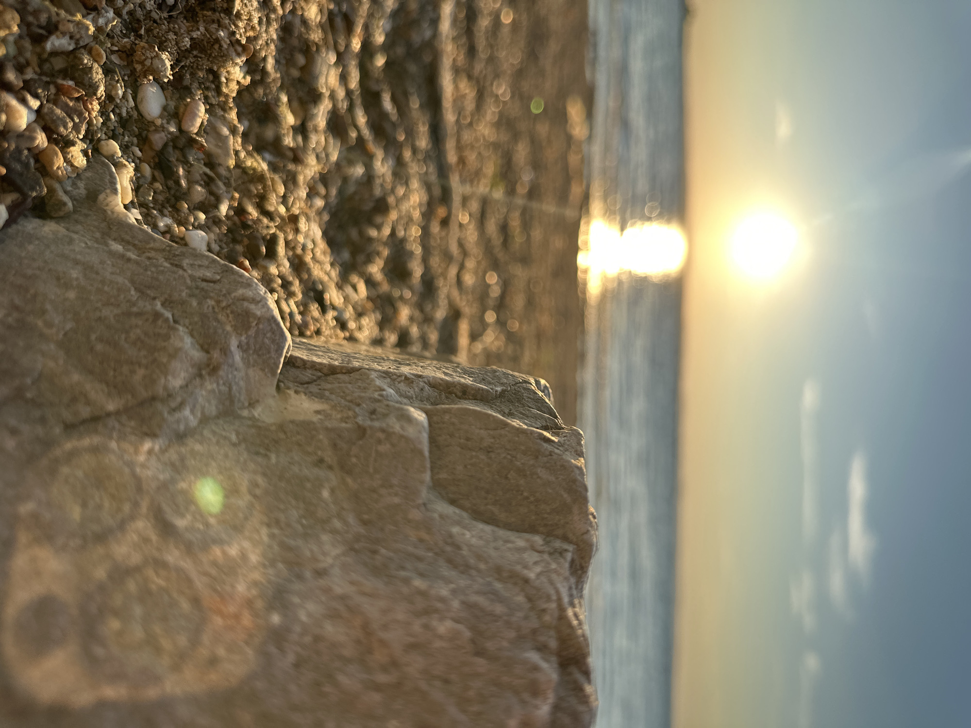 An extreme close-up, low-angle shot captures a large, light-colored rock in the foreground, positioned to the right of the frame. The rock's textured surface is illuminated by the warm glow of the setting sun. Beyond the rock, a beach of pebbles and sand stretches towards the ocean, with the water's surface reflecting the sun's light. The sun itself is a bright, prominent orb in the upper left portion of the image, casting a strong reflection on the water. The sky transitions from a pale blue at the top to a warm yellow-orange hue near the horizon. The background is slightly blurred, suggesting a shallow depth of field. There are no people or specific structures identifiable in the image, creating a serene and natural beach scene. The location is Energetik, Russia.