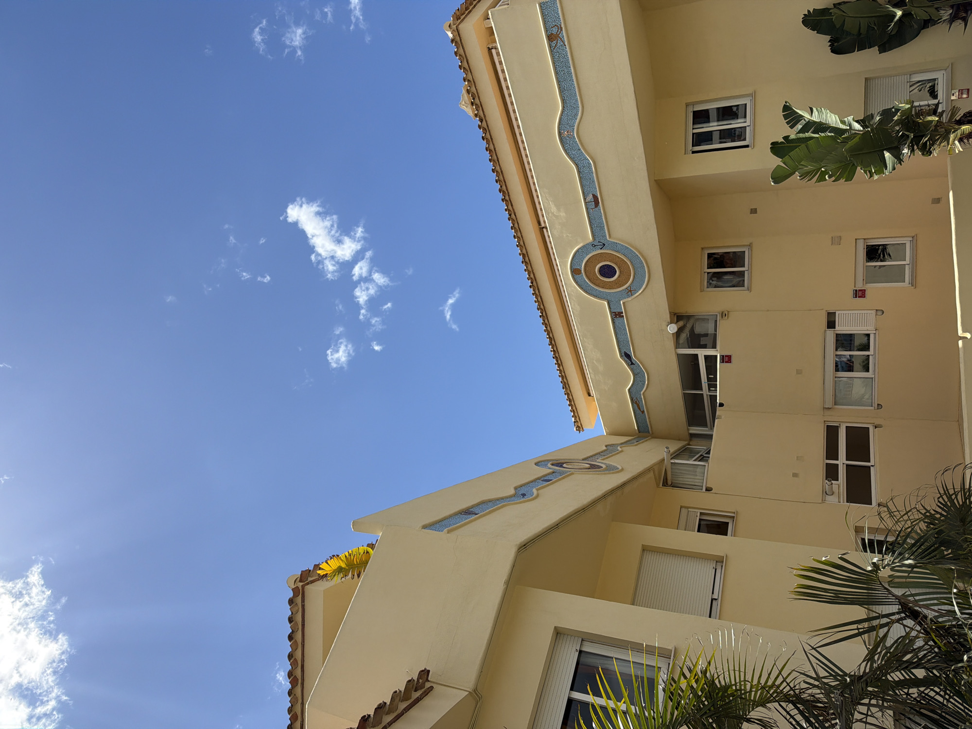 The photograph shows a low-angle view of a multi-story building in Benalmádena, Spain, set against a bright blue sky with scattered white clouds. The building is painted a light yellow color, and features a unique decorative design on its facade: a flowing, light blue mosaic pattern that runs vertically. This design includes circular motifs. The building has multiple windows, some with white blinds, arranged in a stacked pattern. Palm trees and other tropical plants are visible near the base of the building.