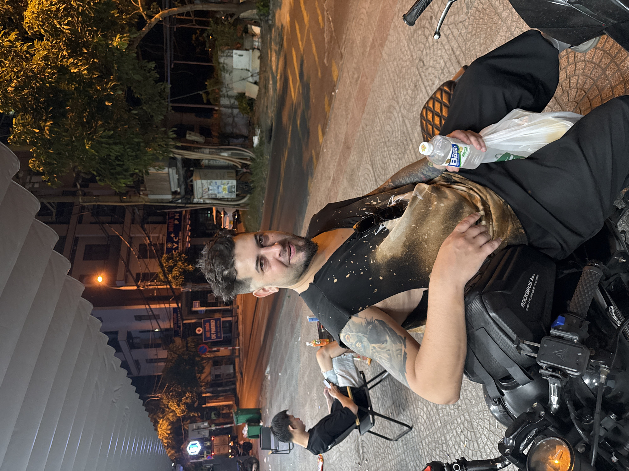 A man is sitting on a motorcycle in Đà Nẵng, Vietnam. He has dark hair, a beard, and tattoos on his arms. He's wearing a black t-shirt with bleach spots and black pants. He is holding a bottle of water and a plastic bag. A man wearing a black shirt sits in a chair behind him, looking down at his phone. There are buildings and trees in the background. Street lights illuminate the scene.