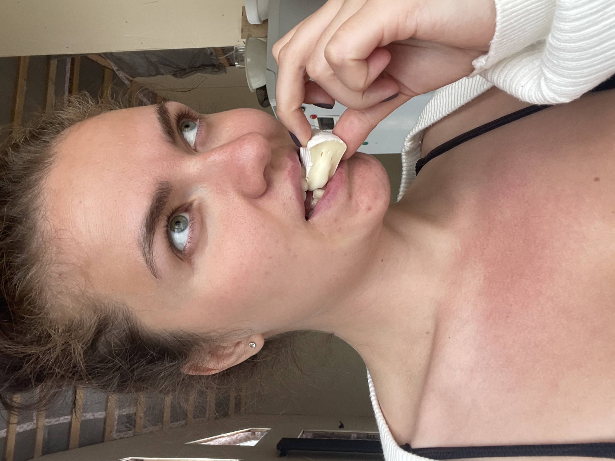 A woman with light skin, green eyes, and light brown hair is holding a piece of Brie cheese to her mouth. She is wearing a white sweater over a black top. The background is blurry, but appears to be an unfinished room with exposed wooden framing.