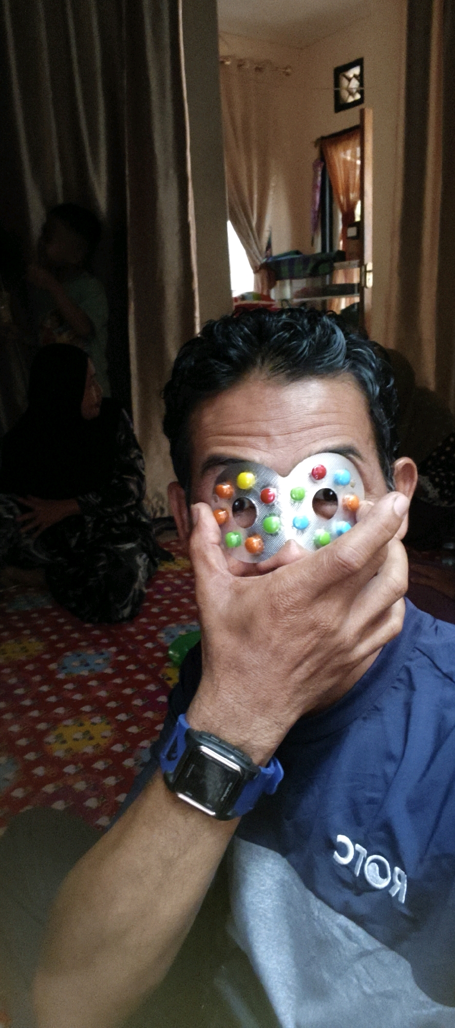 In Muaro Jambi, Indonesia, a man is holding a colorful candy-filled novelty item over his eyes, obscuring his face. The man, with dark, curly hair, wears a blue collared shirt with white lettering on the chest and a blue digital watch on his left wrist. His right hand is raised to his face, holding the candy item. In the background, partially visible through curtains, are two women and a child. The setting appears to be indoors, with a patterned red and yellow carpet on the floor and a wall with a window and a framed picture.