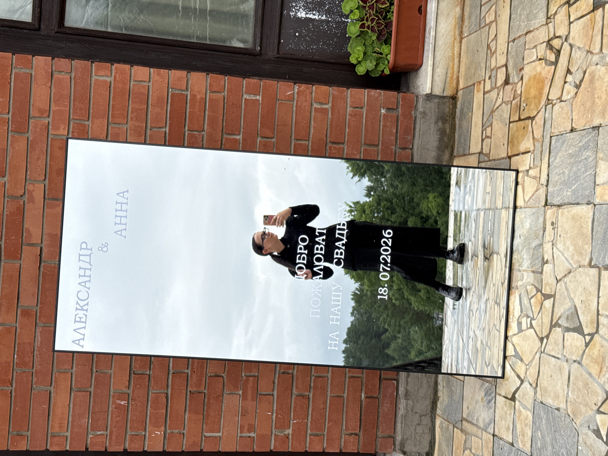 The image shows a full-length mirror standing vertically outside a building. The mirror reflects a person, who appears to be a woman, taking a selfie with a mobile phone. The mirror is inscribed with text in Russian. The top of the mirror displays "АЛЕКСАНДР & АННА". Below this, centered, are the words "ДОБРО ПОЖАЛОВАТЬ НА НАШУ СВАДЬБУ" (Welcome to our wedding). At the bottom of the mirror is the date "18. 07.2026". The background reflected in the mirror shows trees and a cloudy sky. The mirror is placed on a paved area with large, irregular stone tiles. Behind the mirror, part of a brick building is visible. The setting appears to be an outdoor area, possibly a courtyard or patio, near a building constructed with red brick. The overall scene suggests an outdoor wedding reception or ceremony. The location is identified as Zavod Mikhailovskii, Russia.